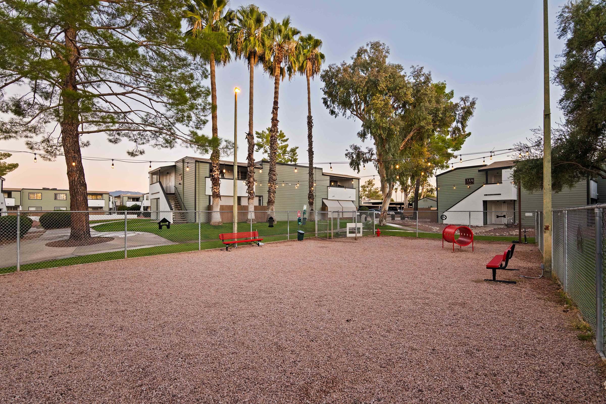 A playground area surrounded by gravel, featuring a red play structure and a couple of benches. Tall palm trees and other greenery are visible in the background, along with low-rise apartment-style buildings. The scene is calm, capturing a sunset ambiance.