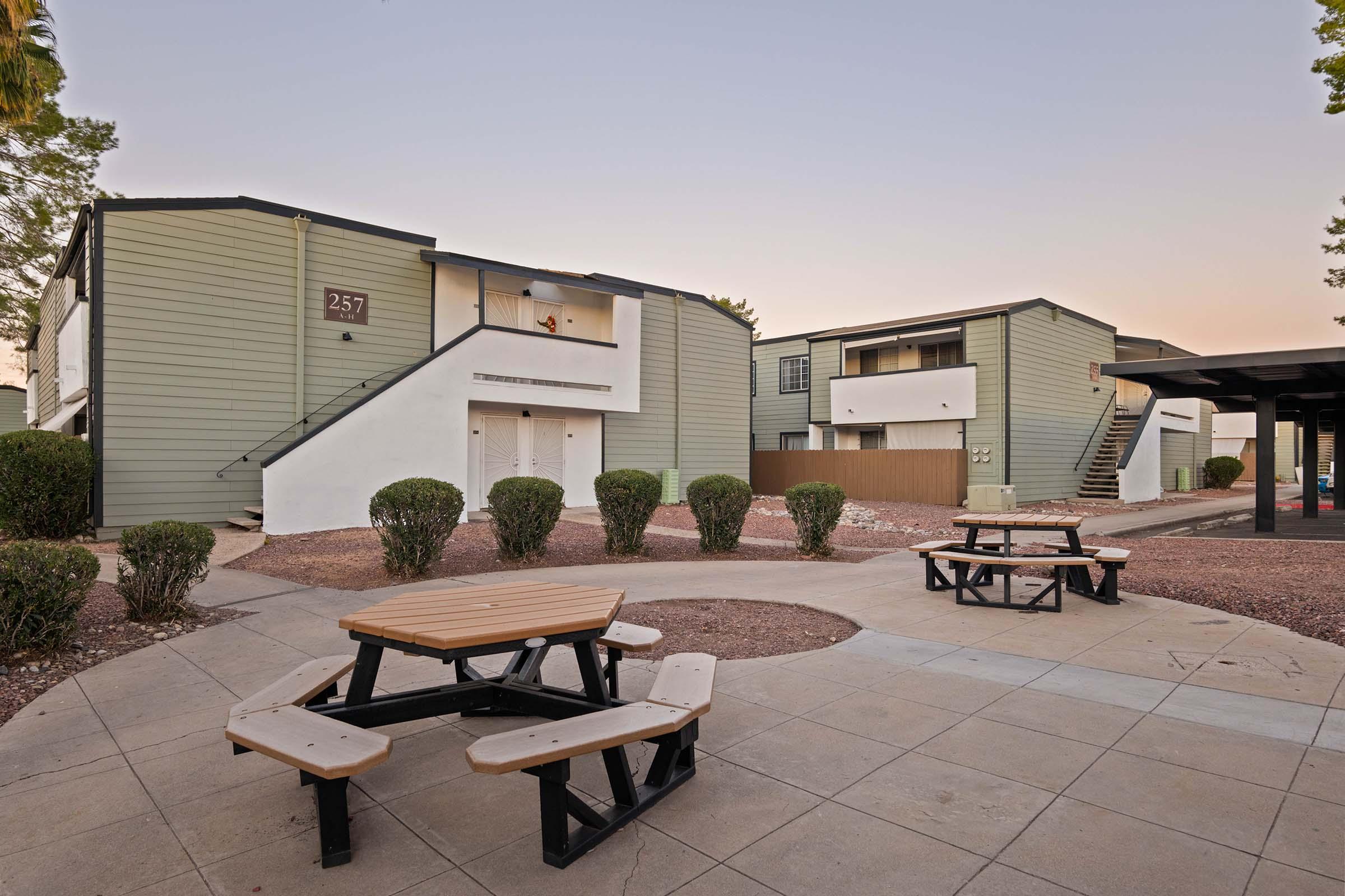 A quiet outdoor area featuring a wooden picnic table surrounded by landscaped shrubs, with two multi-story apartment buildings in the background. The buildings have a modern design with light green siding and white accents, and the scene is illuminated by the soft light of early evening.