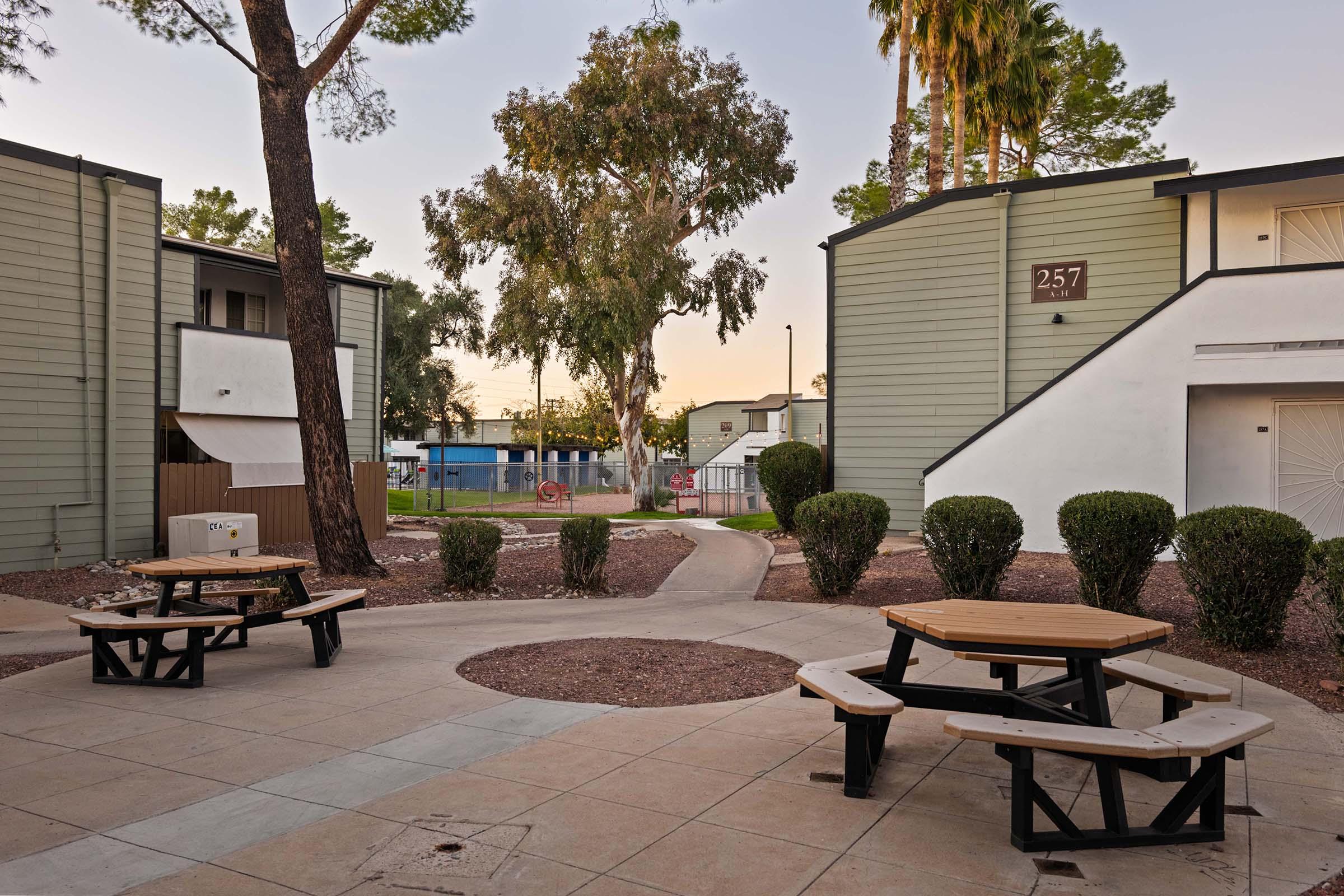 A peaceful outdoor courtyard featuring multiple picnic tables surrounded by well-maintained shrubbery. The scene includes two buildings with light green exteriors and a pathway leading towards a fenced area in the background. Soft lighting suggests early evening, with trees providing shade.