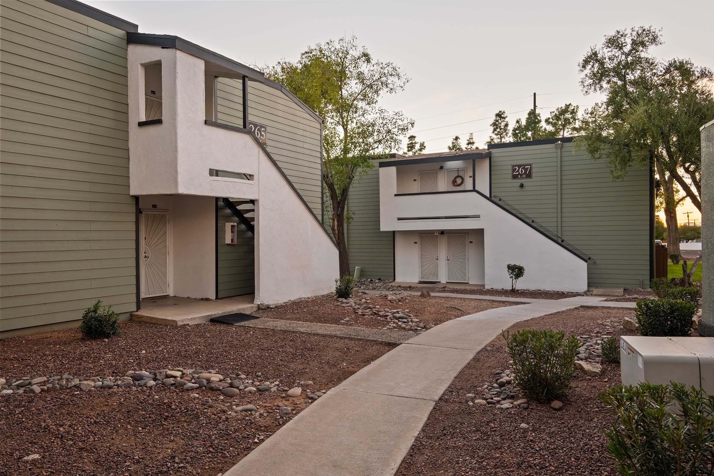 A view of a modern apartment complex featuring two buildings with a minimalist design. The buildings have green and white exteriors, surrounded by landscaped pathways, rocks, and small shrubs. The scene is set during the early evening with soft lighting.