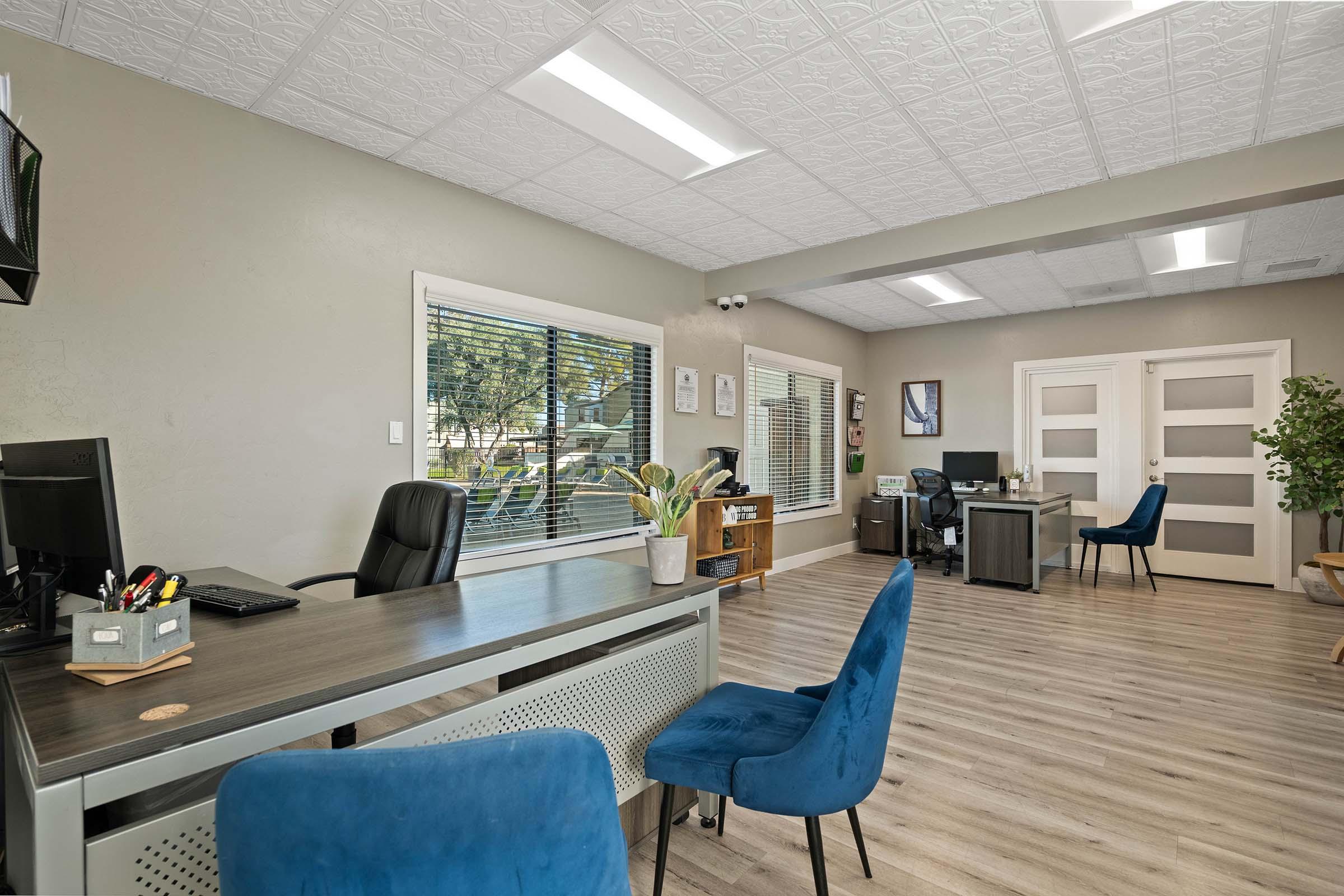 Interior of a modern office space featuring a desk with a computer, stylish blue chairs, large windows providing natural light, and contemporary decor. The room has wooden flooring and a light-colored ceiling with a patterned design. There are plants and organizational elements on display.