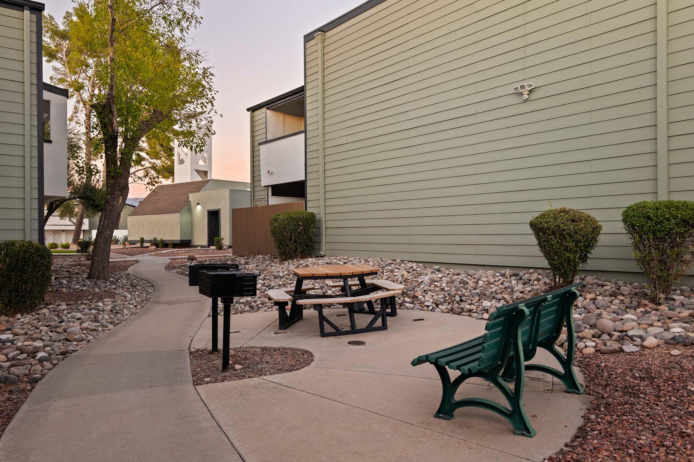 A small outdoor area featuring a circular path, a picnic table with benches, and a charcoal grill surrounded by landscaping with rocks and shrubs. Nearby buildings are visible in the background, along with greenery and a clear sky at dusk.