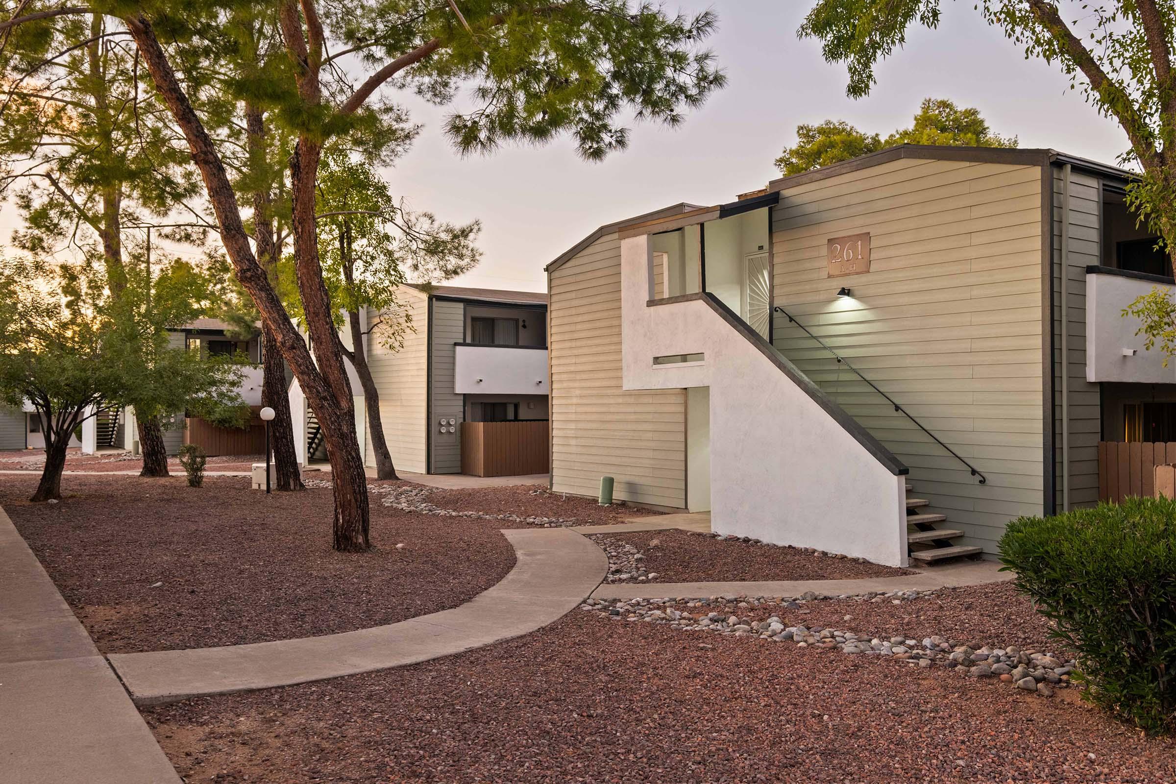 A modern apartment complex with wooden exterior and a staircase leading to the entrance. The building is surrounded by trees and a pathway, creating a tranquil outdoor scene. The setting is well-maintained, with gravel landscaping and lush greenery. The image captures a peaceful residential environment.