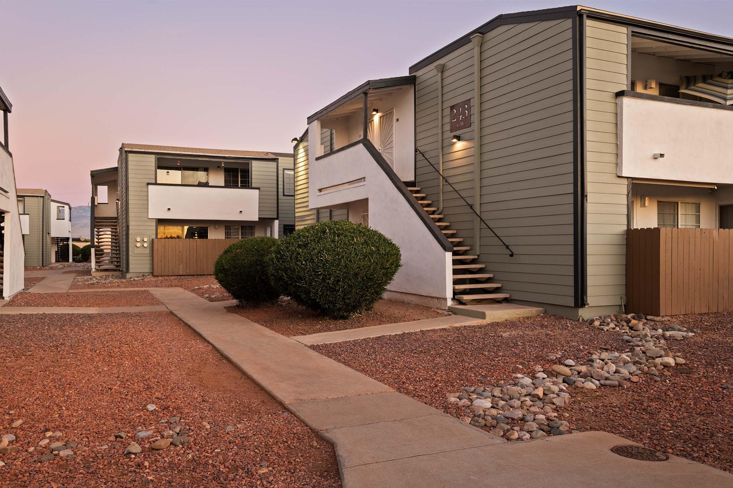 A quiet residential area featuring two-story apartment buildings. The path leads to a staircase, surrounded by landscaped bushes and gravel. The soft evening light creates a warm atmosphere, highlighting the architecture and outdoor space.