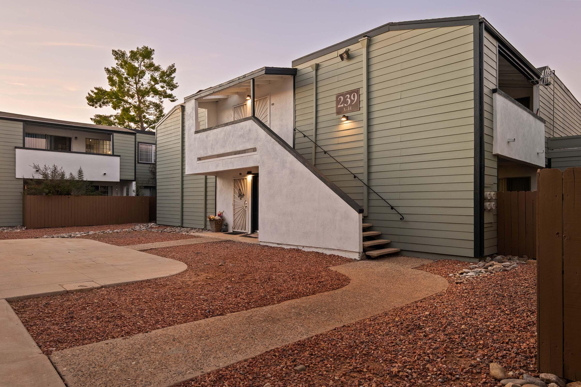 A modern two-story building with a prominent staircase and a landscaped yard featuring gravel pathways. The exterior has a mix of green and white siding. The scene is set in a quiet area with nearby structures visible in the background, illuminated by soft evening light.