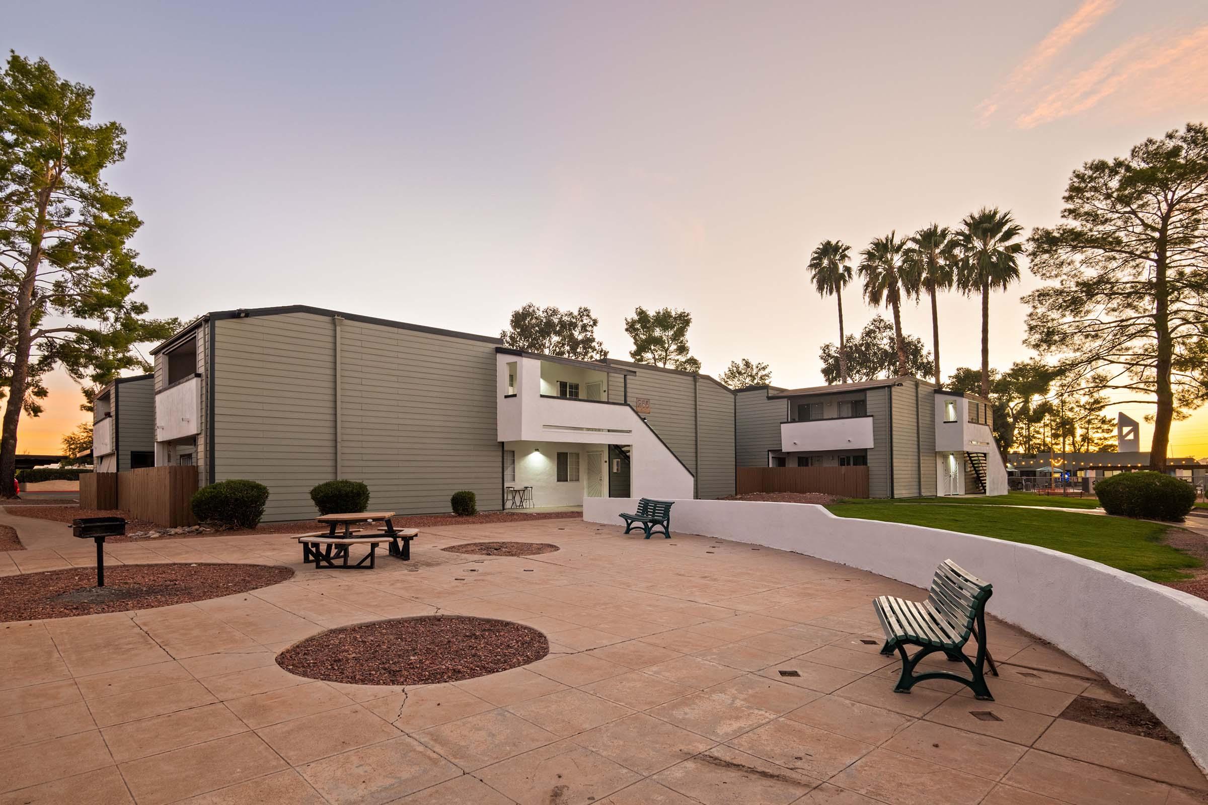 A peaceful outdoor area featuring two-story green apartment buildings, with palm trees in the background under a colorful sunset sky. A picnic table and benches are placed on a paved surface, surrounded by low shrubs, creating a welcoming community space.