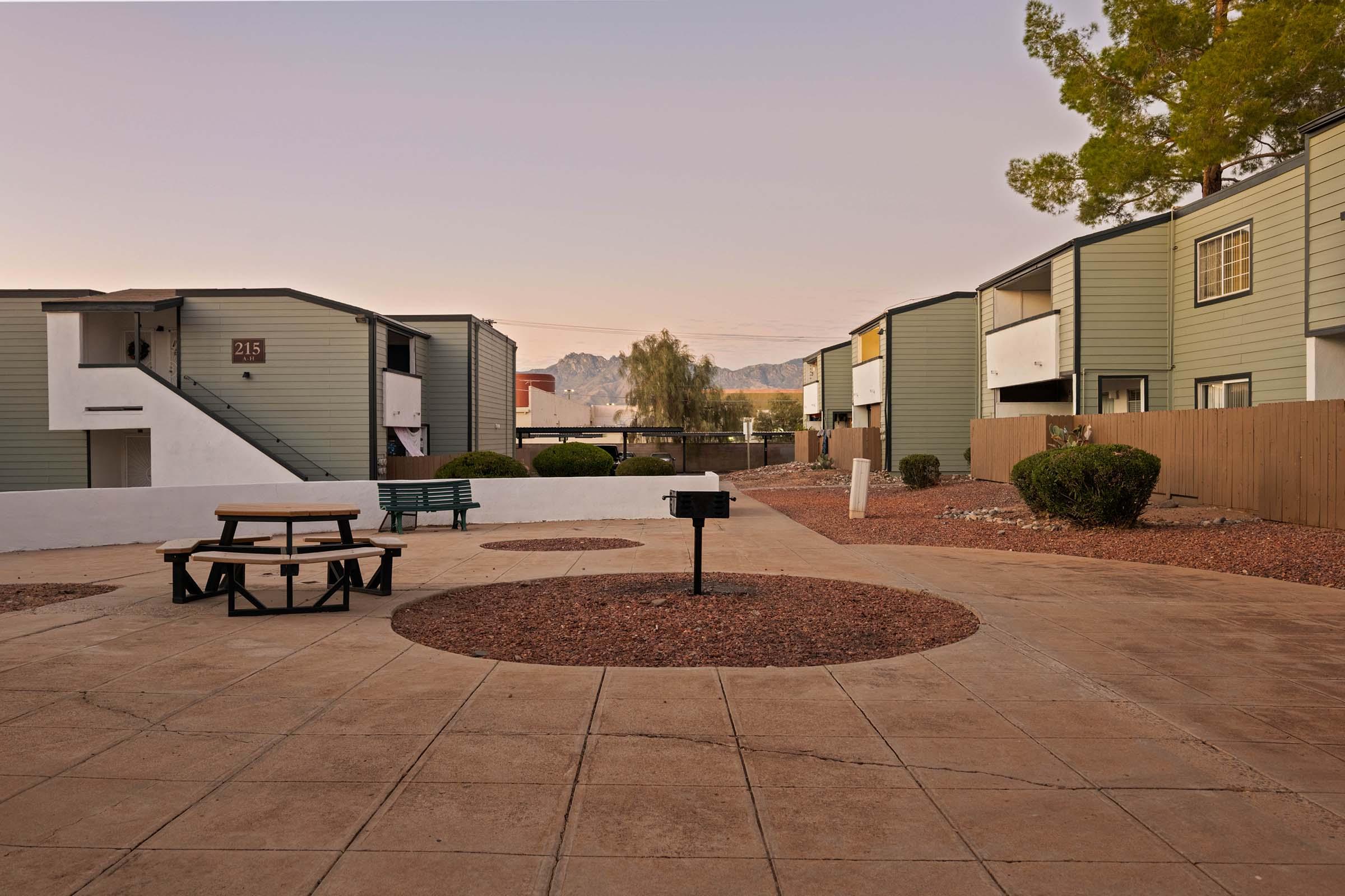 View of a residential courtyard with several two-story apartment buildings on either side. A picnic table sits in the center on a circular concrete area, surrounded by gravel and small shrubs. Trees are visible in the background, and the sky shows hints of pink as the sun sets.