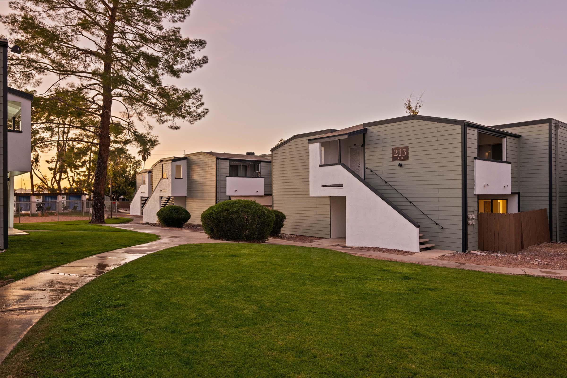 A landscaped pathway leads through a residential complex featuring modern, two-story apartment buildings. The structures have a mix of light green and white exteriors, with stairs leading to the upper levels. Surrounding the buildings are well-maintained lawns and tall trees, with a soft sunset glow in the background.