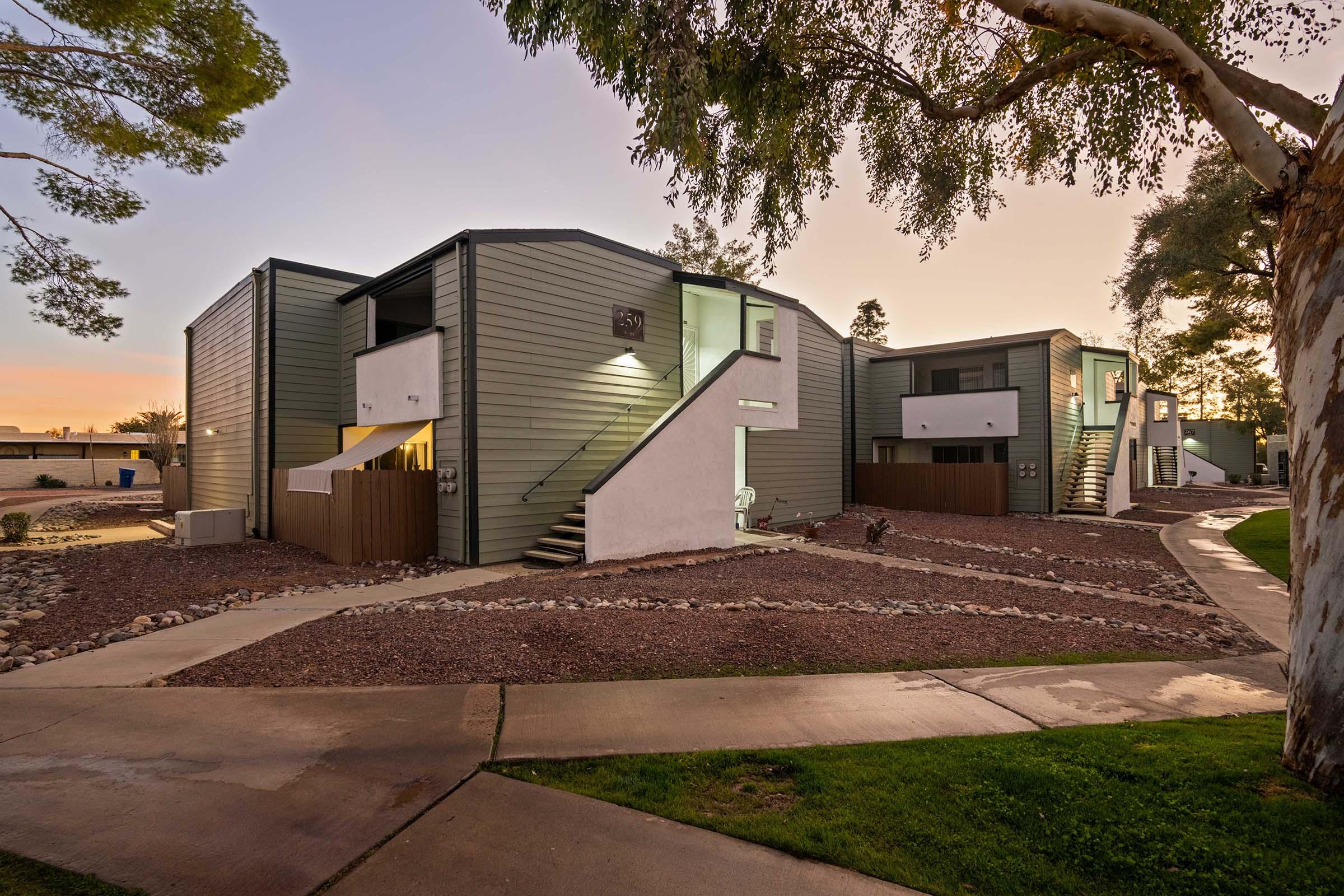 Two modern multi-story apartment buildings with a gray exterior, surrounded by landscaped pathways and trees. The area features a mixture of gravel and grass, with well-maintained walkways leading to the entrances of the apartments. The scene is set during twilight, with soft lighting.