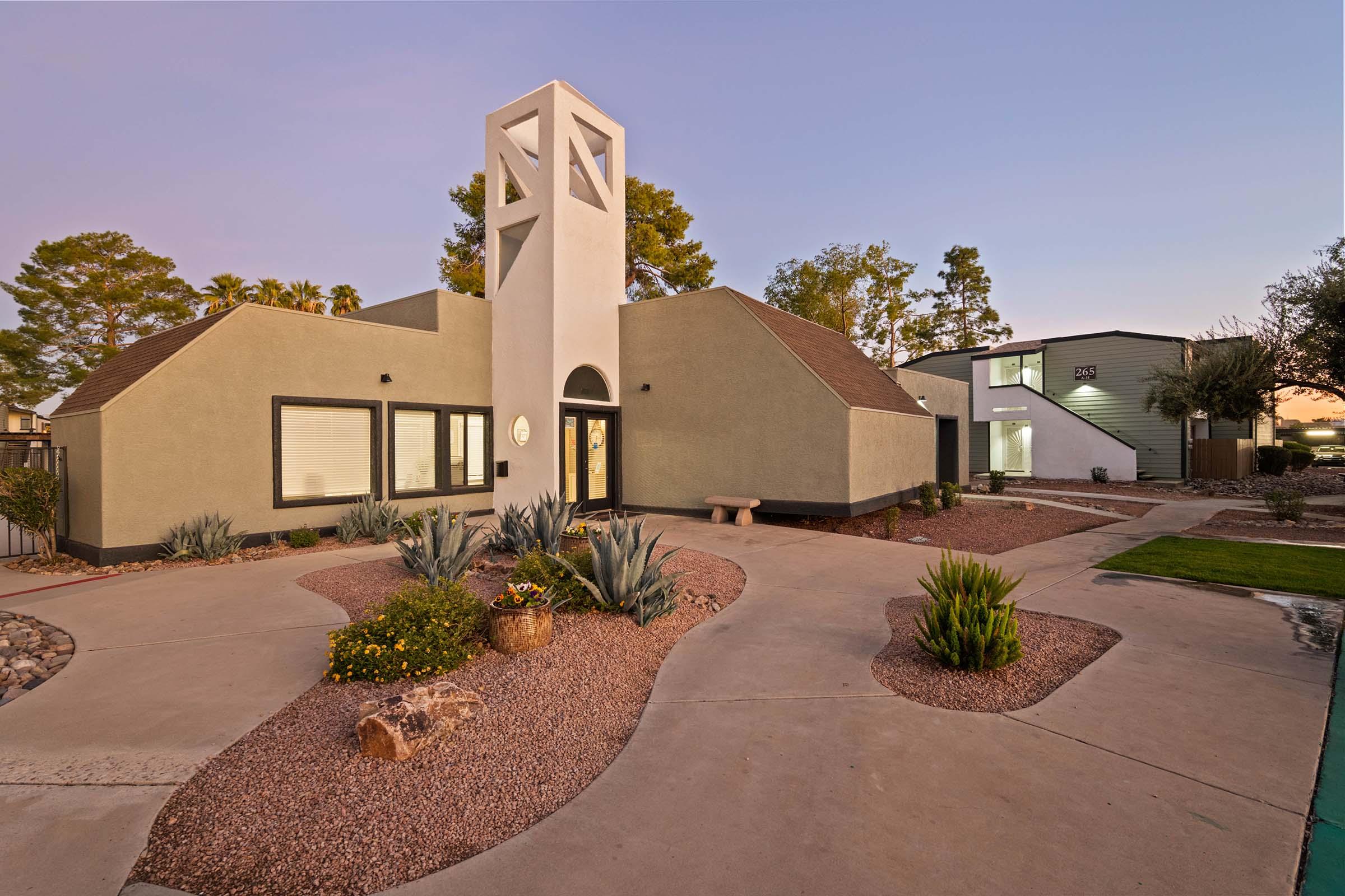 A modern house with a distinctive tower and landscaped garden featuring desert plants, rocks, and a stone bench. The pathway leads to the entrance, surrounded by greenery and flower beds, set against a backdrop of trees under a clear sky during twilight.