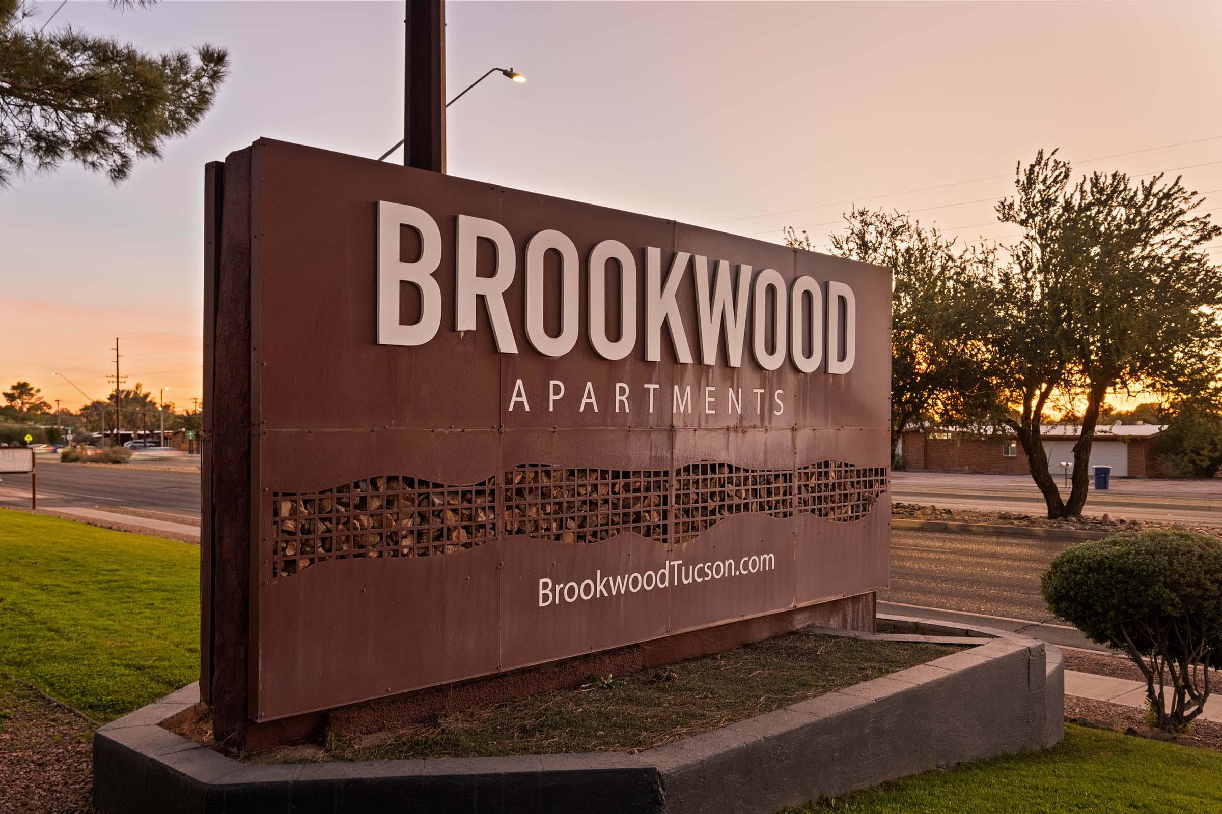 Sign for Brookwood Apartments with the website URL "BrookwoodTucson.com" displayed. The sign is set against a sunset sky, surrounded by greenery and a well-maintained landscape.