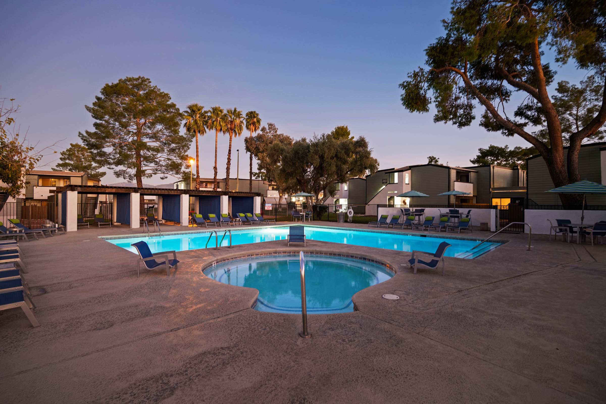 A serene evening view of a swimming pool area featuring a main pool and a connected hot tub. Surrounded by lounge chairs, trees, and nearby buildings, the scene is illuminated by soft lighting, creating a relaxing atmosphere. Palm trees dot the background against a twilight sky.