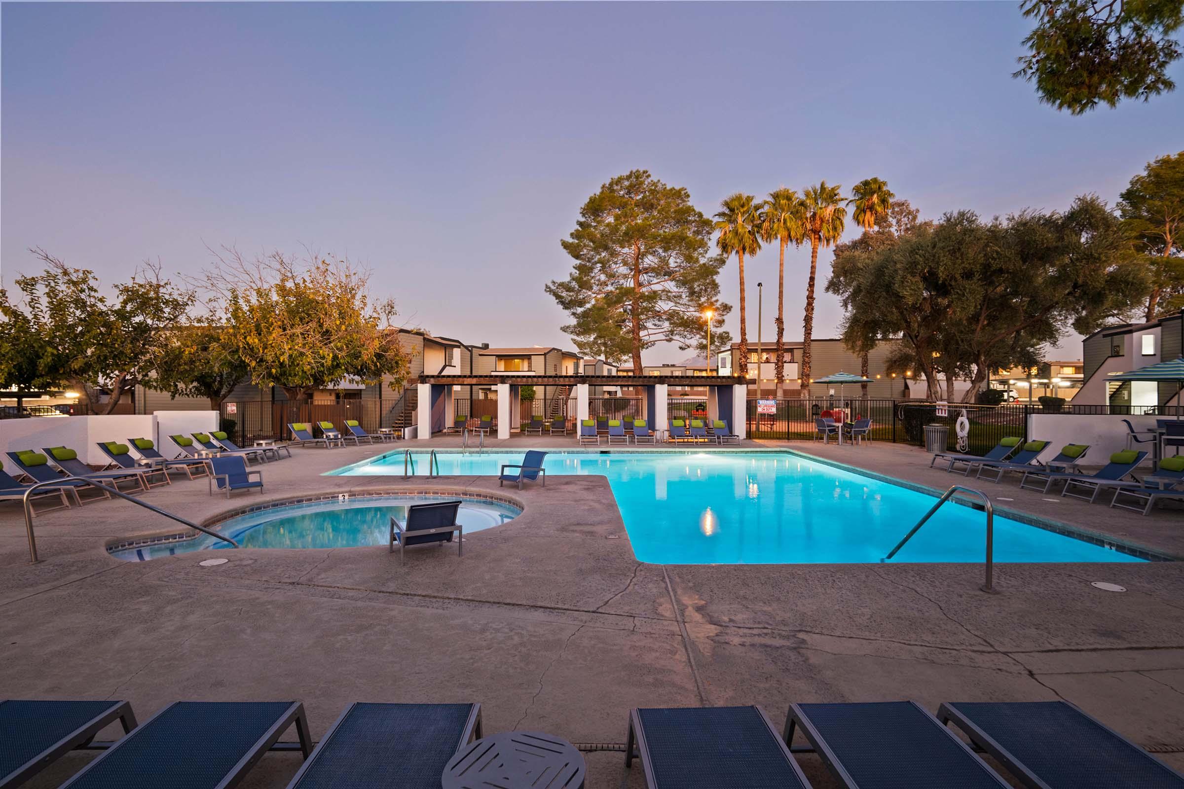 A serene outdoor pool area in the evening, featuring a large swimming pool with bright blue water. Comfortable lounge chairs surround the pool, and a hot tub is visible nearby. Lush palm trees and greenery provide a relaxing ambiance, and cozy lighting enhances the inviting atmosphere.