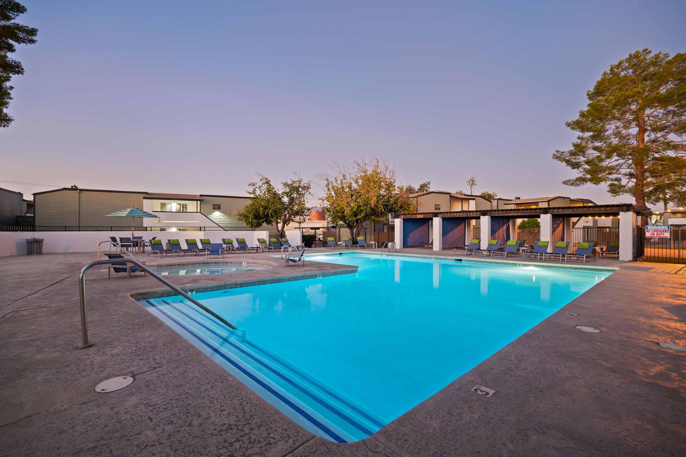 A sparkling blue swimming pool surrounded by lounge chairs and shaded areas. The scene is set in the late afternoon or early evening, with a clear sky and trees providing some greenery. The pool area is well-maintained, inviting for relaxation and leisure activities.