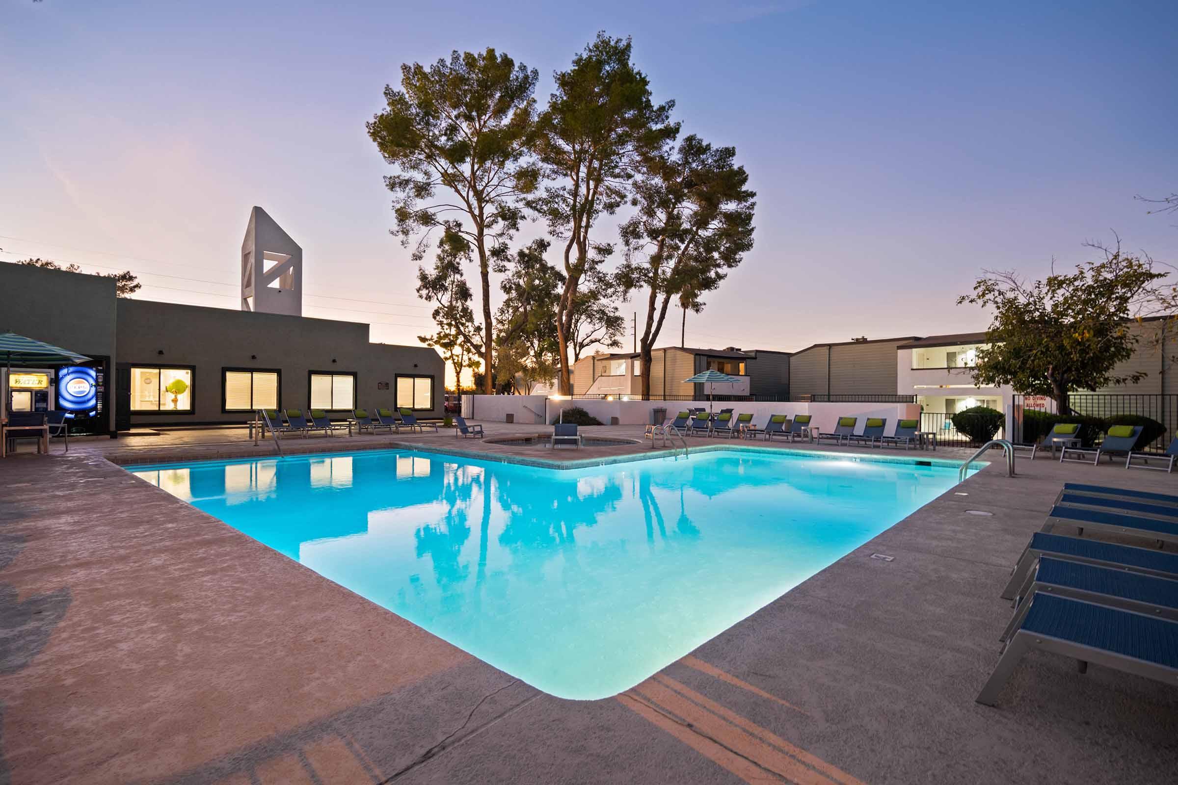 A serene outdoor swimming pool surrounded by lounge chairs, with tall trees in the background. The pool reflects the soft colors of a sunset sky, creating a relaxing atmosphere. Nearby buildings provide a modern backdrop, and a shaded seating area is visible on one side.