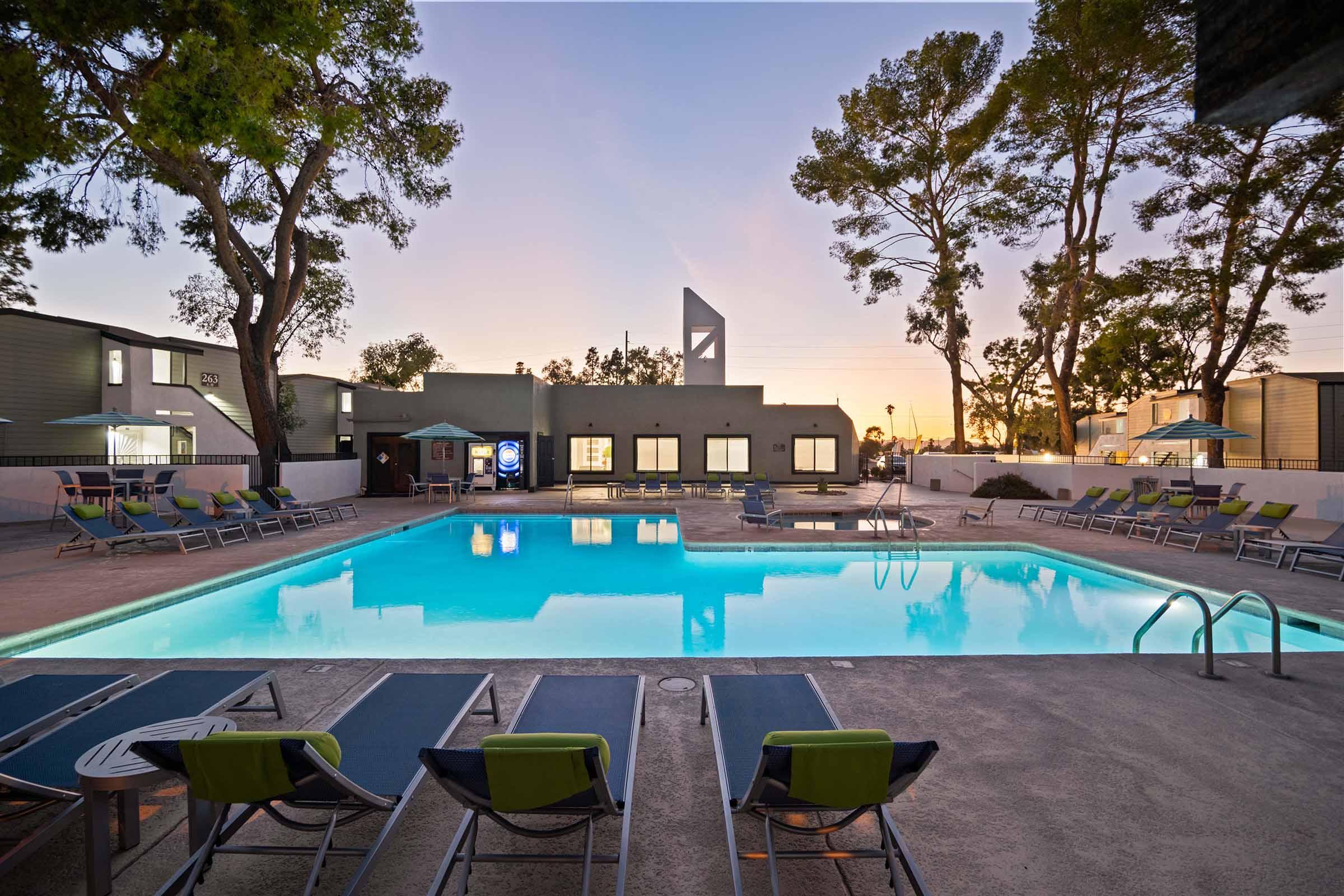 A serene swimming pool with crystal-clear water surrounded by lounge chairs. In the background, trees partially shade the area, and a modern building is visible. The sky is painted in soft pastel colors as the sun sets, creating a tranquil and inviting atmosphere for relaxation.