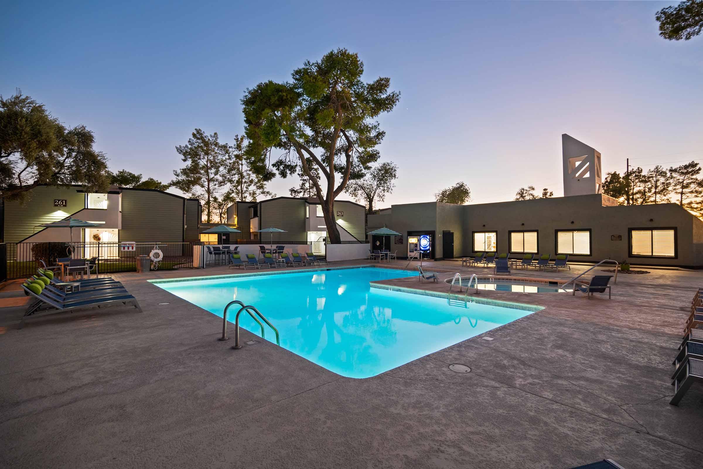A serene swimming pool surrounded by lounge chairs at dusk, with well-maintained buildings and tall trees visible in the background. Soft lighting enhances the tranquil atmosphere, creating a welcoming space for relaxation and leisure.