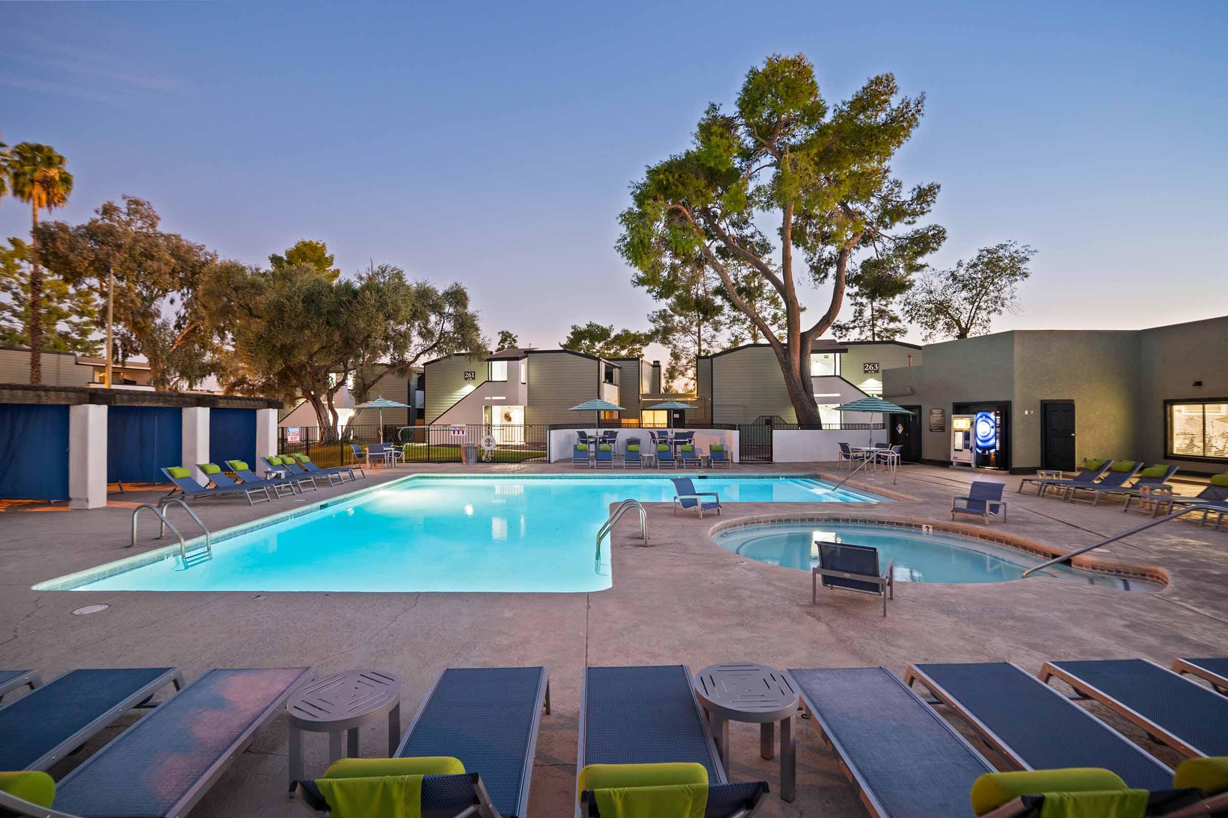 A spacious outdoor pool area surrounded by lounge chairs and a hot tub, with palm trees in the background. The setting is tranquil, featuring a well-maintained pool surrounded by green landscaping and nearby residential buildings. The sky is painted in soft evening hues.
