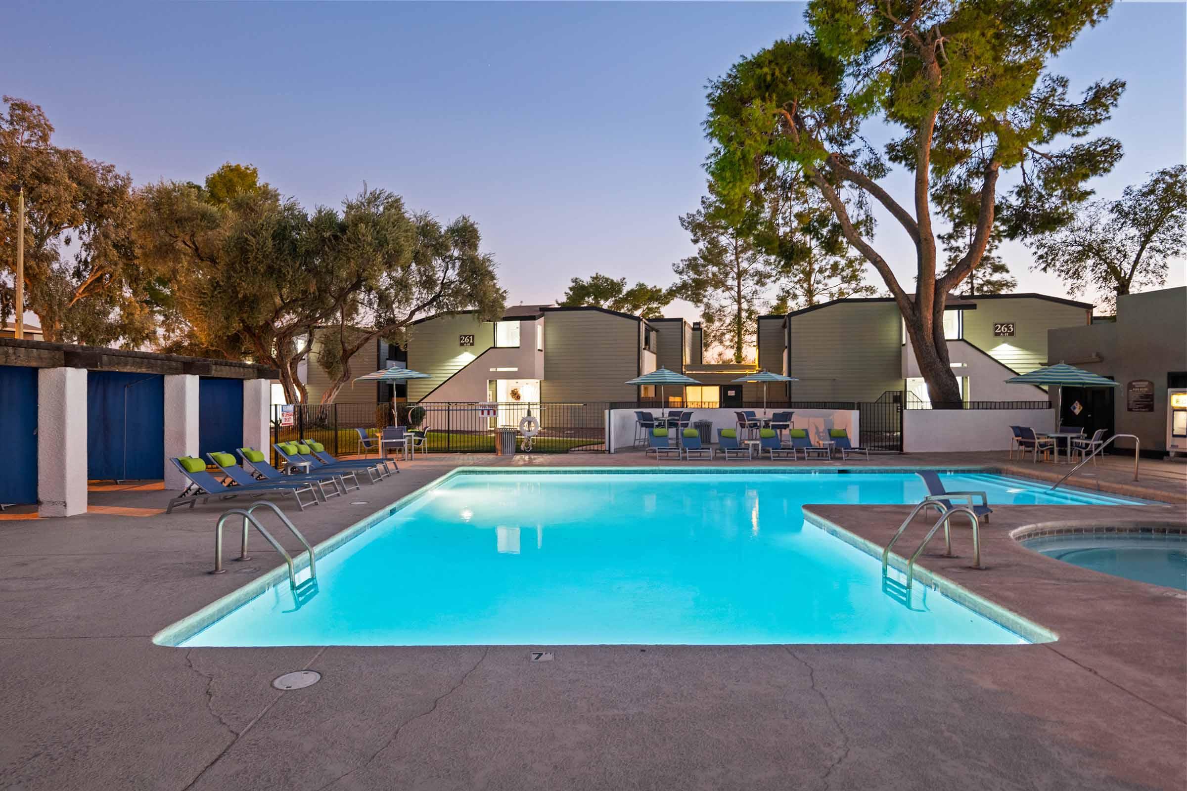 A tranquil swimming pool surrounded by lounge chairs, with a backdrop of trees and residential buildings. The pool is illuminated by soft lighting as evening approaches, creating a calm and inviting atmosphere for relaxation.