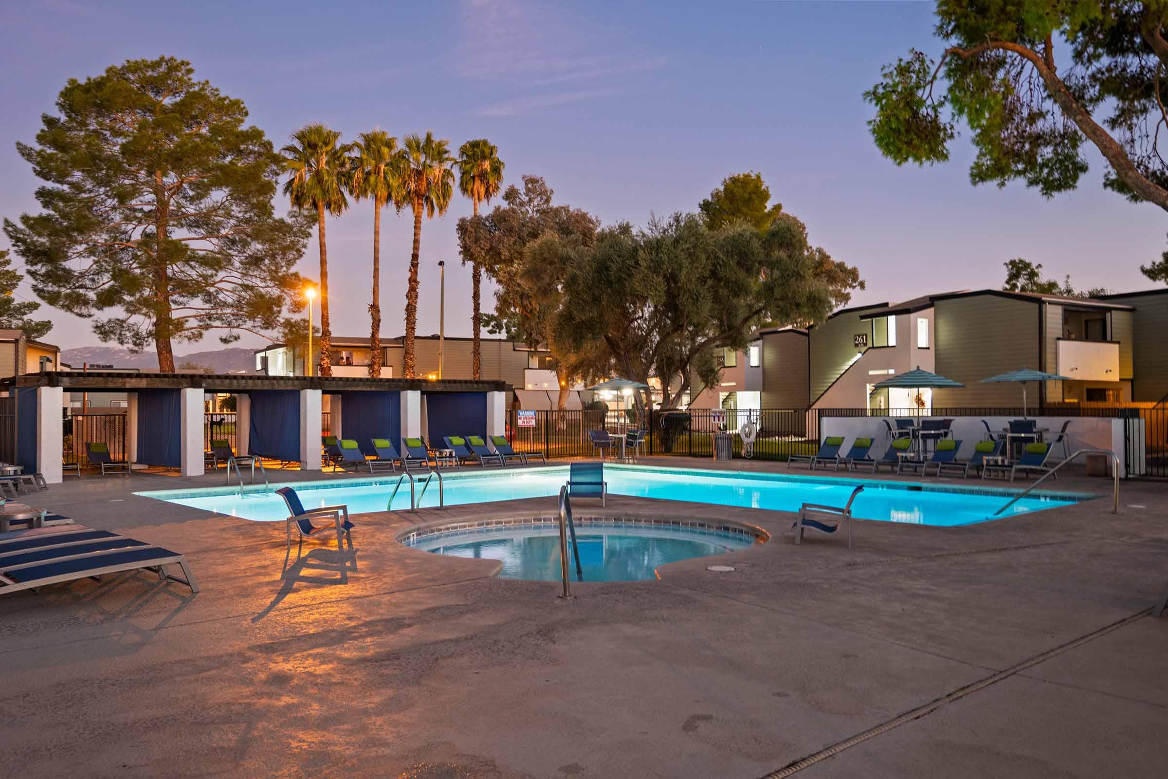 A serene outdoor pool area at dusk, featuring a swimming pool and a hot tub surrounded by lounge chairs. Palm trees and shrubs provide a tropical ambiance, and nearby, a few buildings are illuminated softly. The scene conveys a relaxing atmosphere ideal for leisure and unwinding.