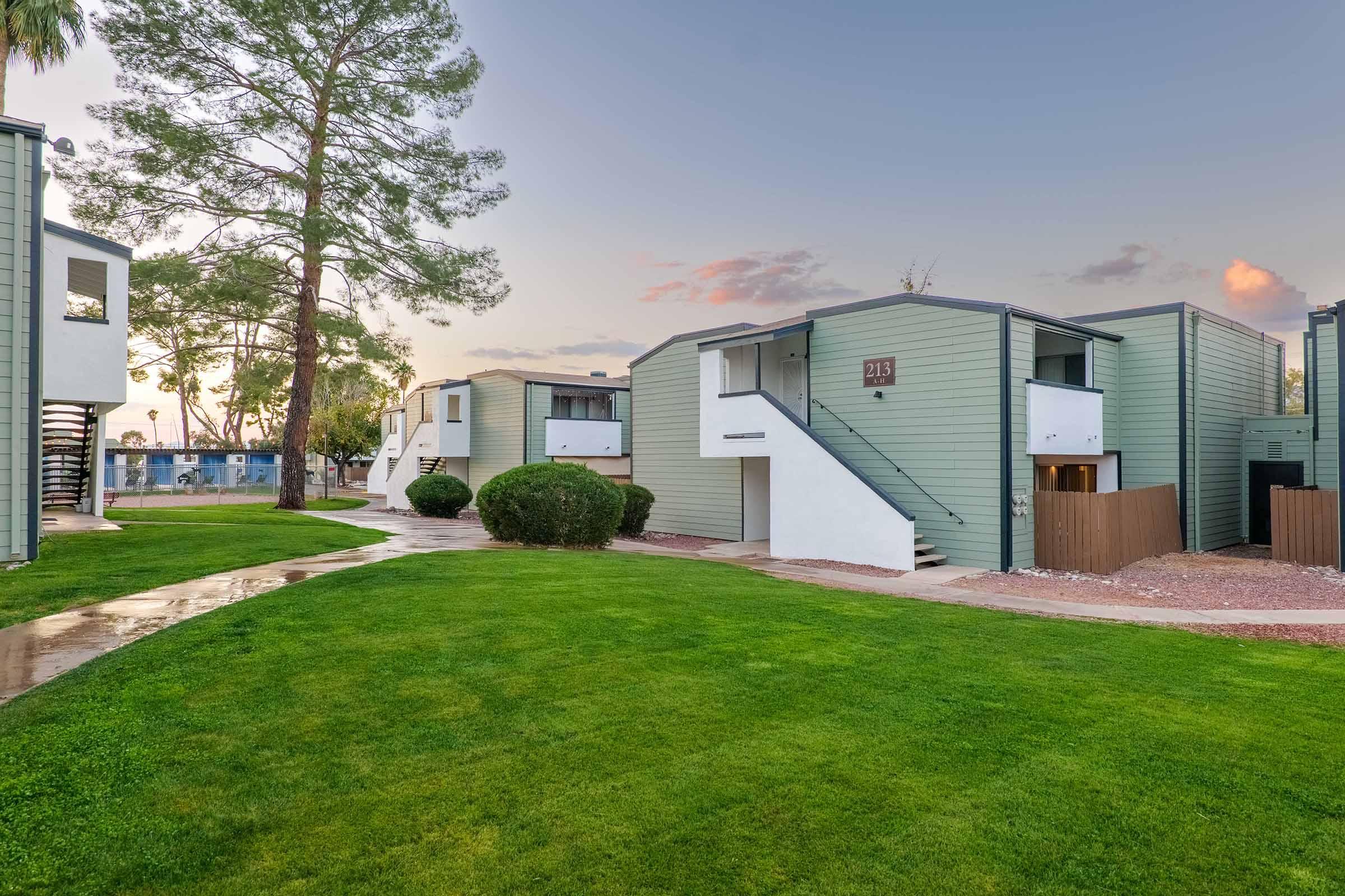 A residential area featuring modern, green-clad duplex buildings surrounded by manicured lawns and trees. The sidewalk leads up to the units, with a soft sunset glow in the background, creating a serene, inviting atmosphere.