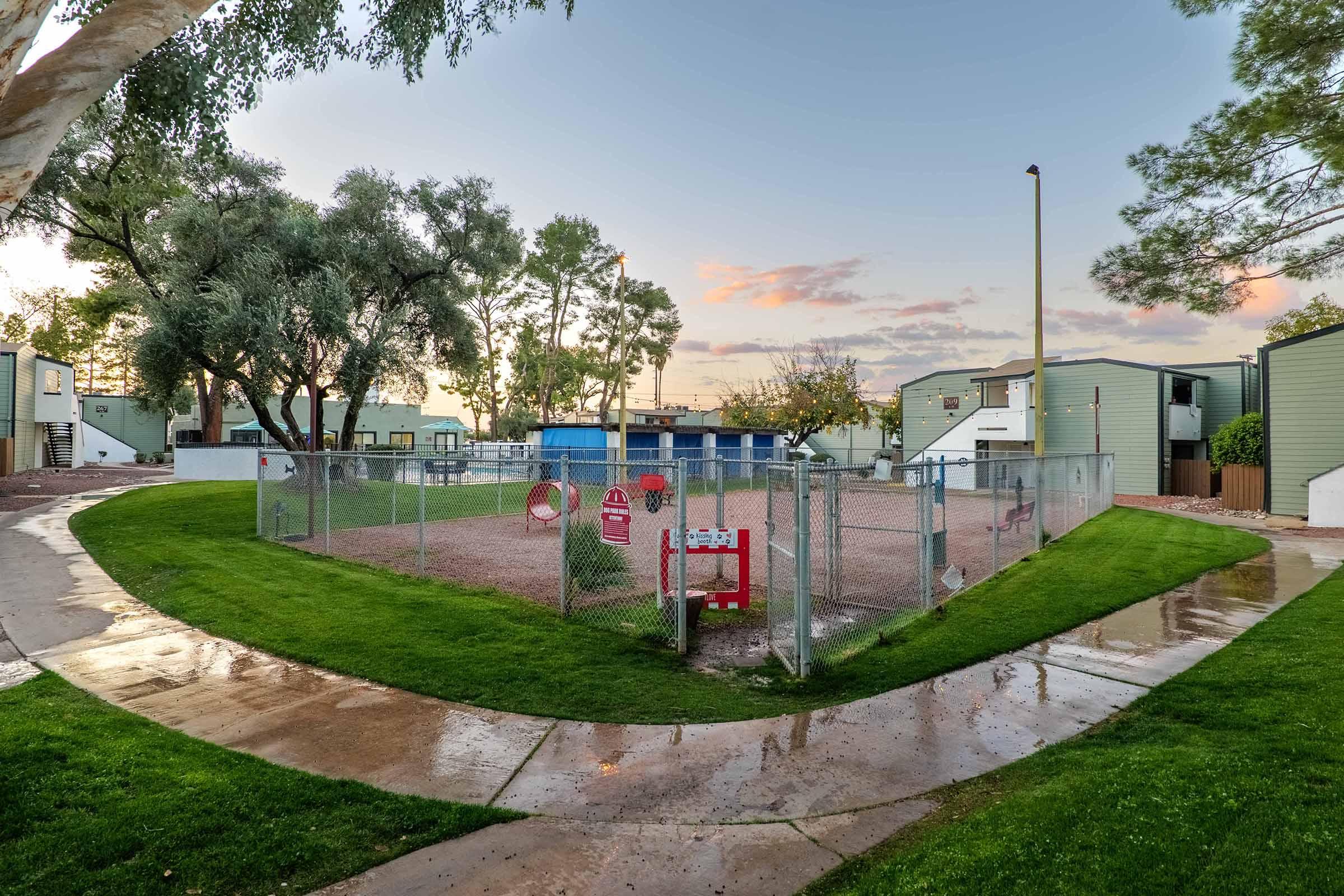 A fenced dog park surrounded by green grass, with a sandy area and a few trees. The park features a water station, dog waste disposal, and benches for pet owners. There are rows of small buildings in the background, hinting at a residential community. The sky has a subtle sunset hue, adding warmth to the scene.