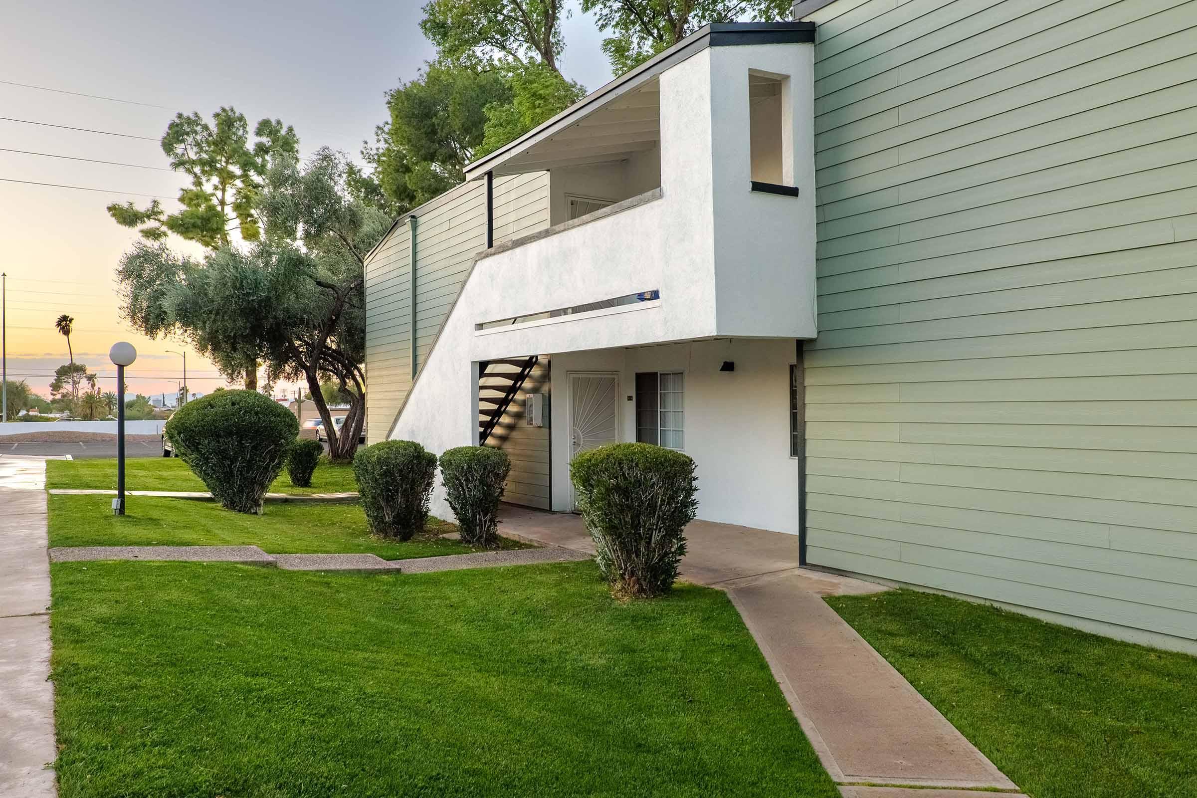 A modern apartment building with green siding, featuring a stairway and a balcony. The landscape includes neatly trimmed shrubs and grass, with a pathway leading to the entrance. There are trees in the background, and the sky shows hints of sunset.