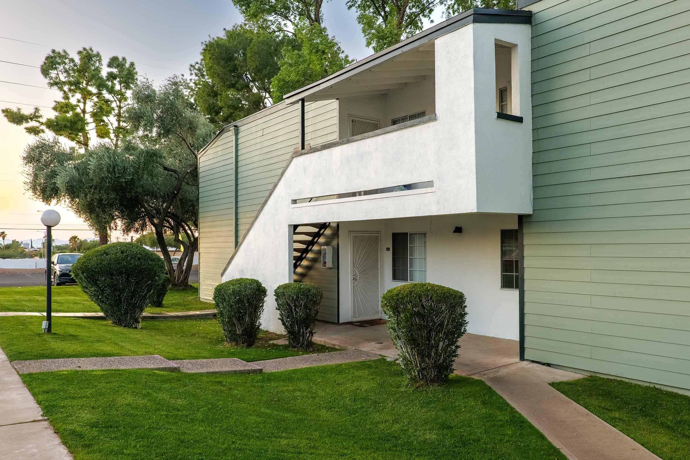A two-story building with a modern, minimalist design. The exterior features light green siding and a white balcony over a stairwell. Surrounding the building are neatly trimmed shrubs and a well-maintained lawn, with a lamp post in the foreground. Trees are visible in the background against a soft evening sky.