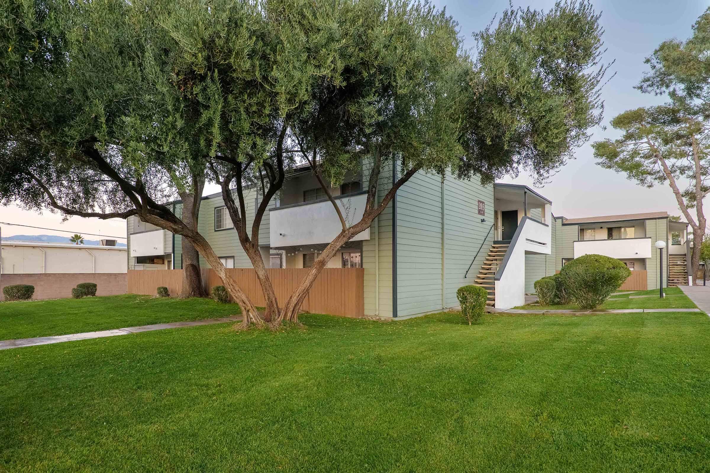 A two-story apartment building with a light green exterior, surrounded by well-maintained grass and shrubs. Two large trees provide shade in the foreground, and there are walking paths visible on the property. The area appears clean and inviting, typical of a residential complex.