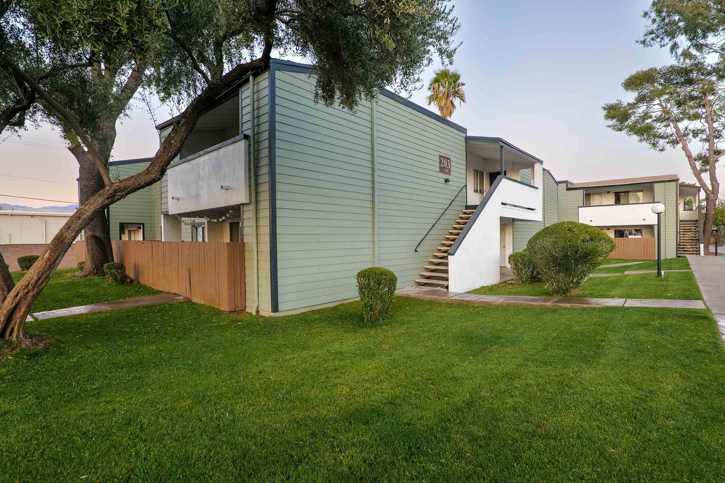 Two-story residential building with a green and gray exterior, featuring a stairway and balconies. Surrounded by well-maintained grass, shrubs, and trees, the scene is at dusk with a calm atmosphere.