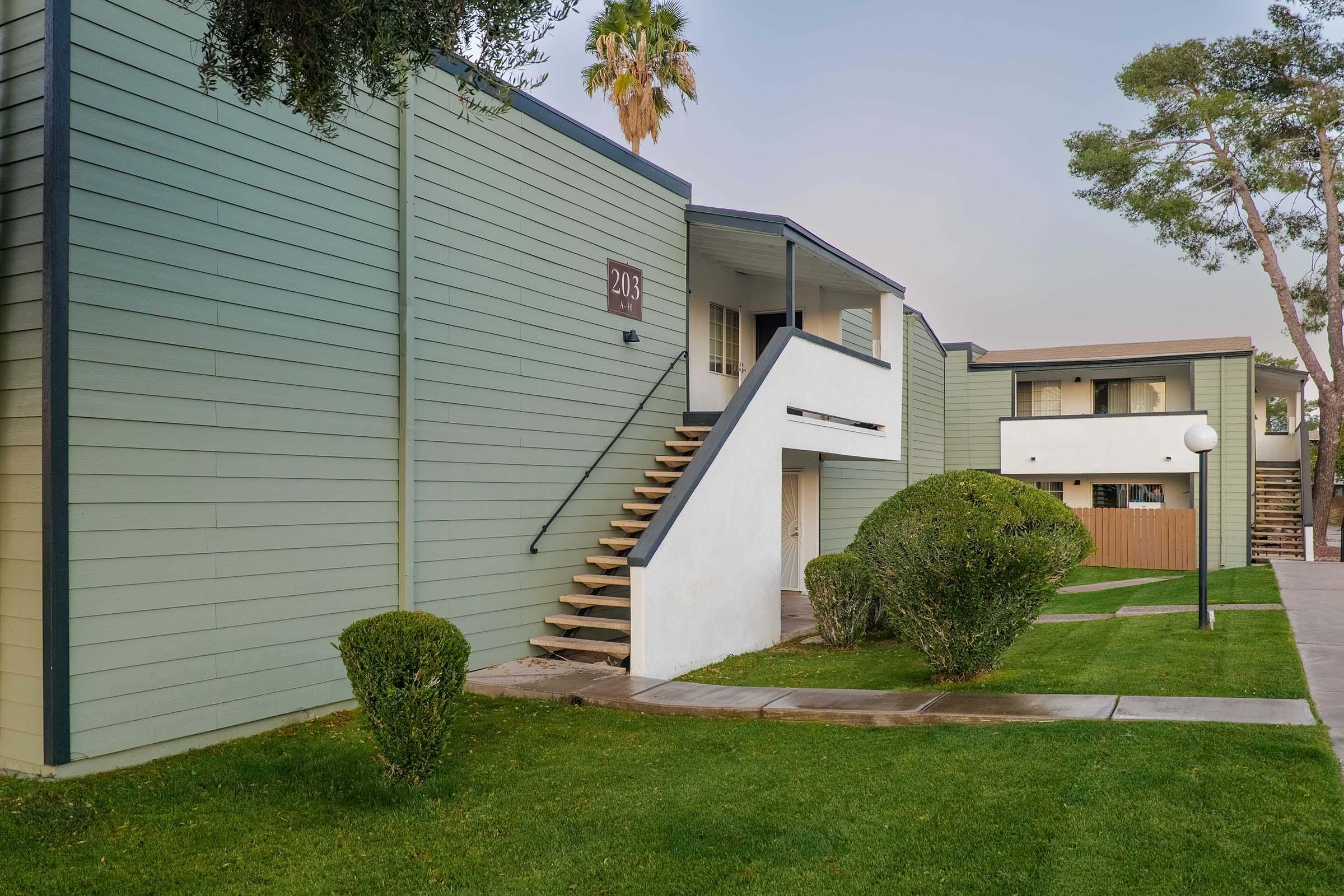 Two-story apartment complex with green exterior walls, featuring a staircase leading to the second floor. The landscape includes neatly trimmed bushes and a grassy area. Palm trees are visible in the background, and pathways provide access to the units. The scene is well-maintained and inviting.