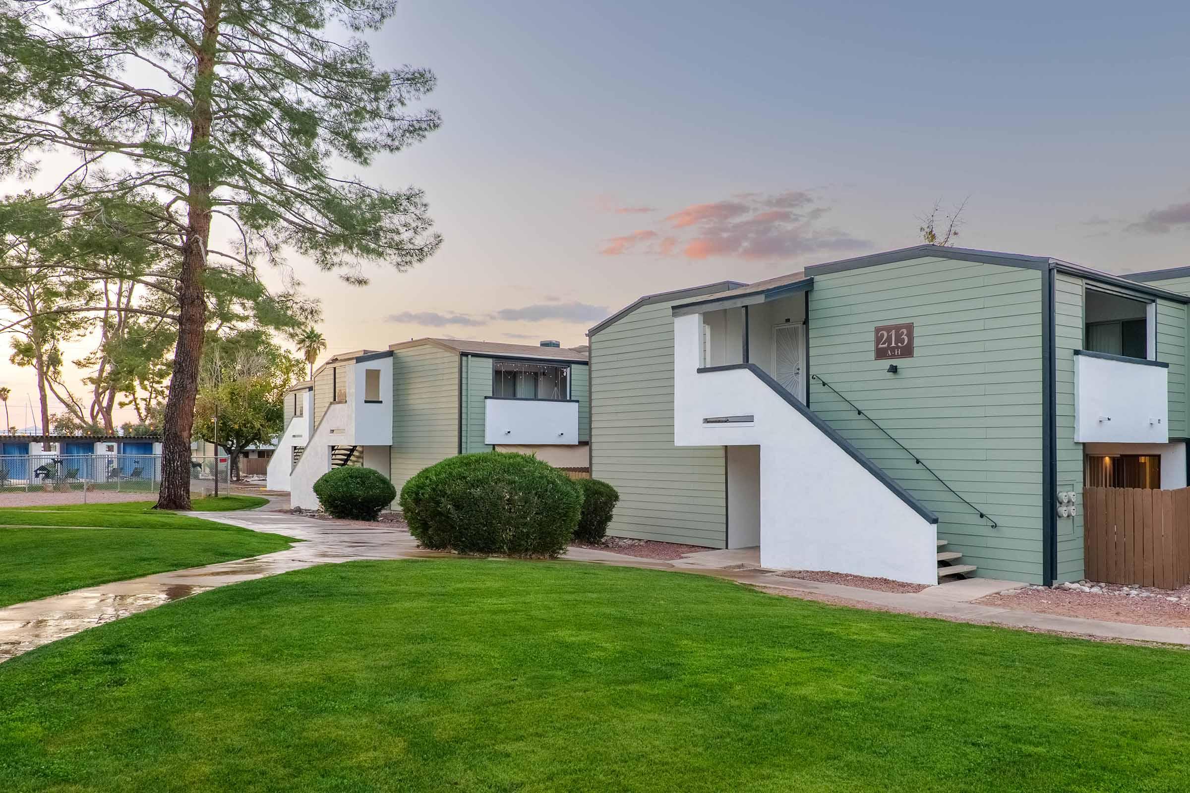 A row of modern, two-story apartment buildings with light green siding, featuring staircases and small balconies. The scene is set in a landscaped area with lush green grass, bordered by shrubs. The sky is slightly overcast, suggesting early evening light.