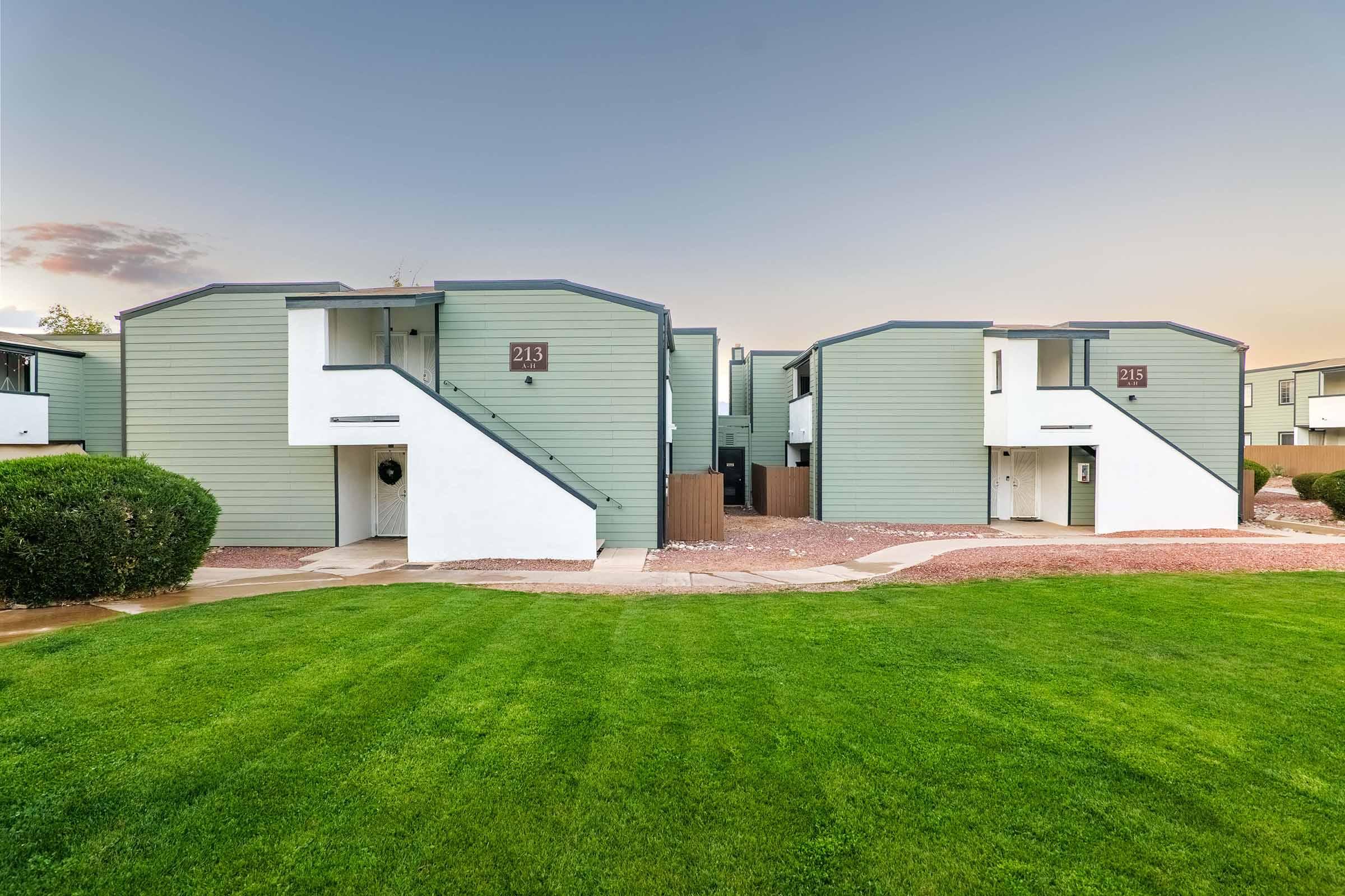 A view of two modern apartment buildings with green exteriors, each featuring a staircase and clearly marked unit numbers. The buildings are surrounded by well-maintained grass and landscaping, with a pathway leading between them. The sky is slightly colorful, indicating either sunrise or sunset.