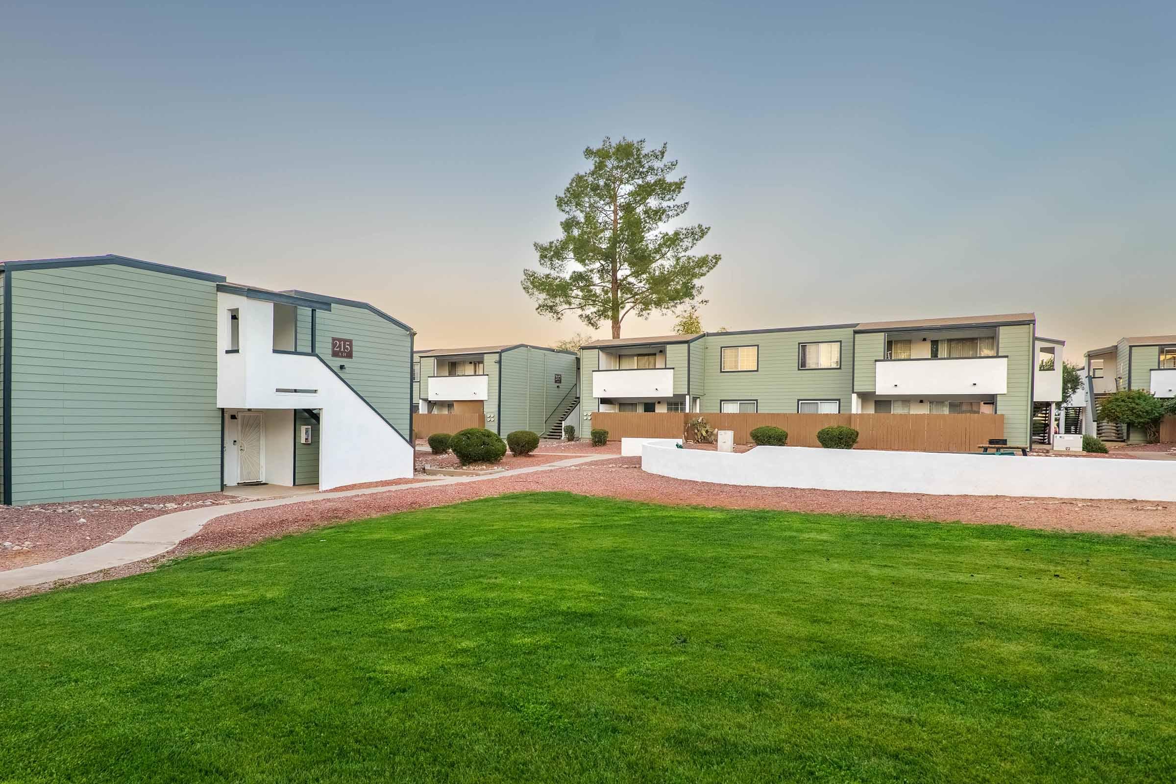 A modern apartment complex featuring two-story buildings with green and white exteriors. The landscape includes well-maintained grass, shrubs, and a pathway leading to the entrance. A large tree stands in the background, under a clear sky at dusk, creating a peaceful ambiance.