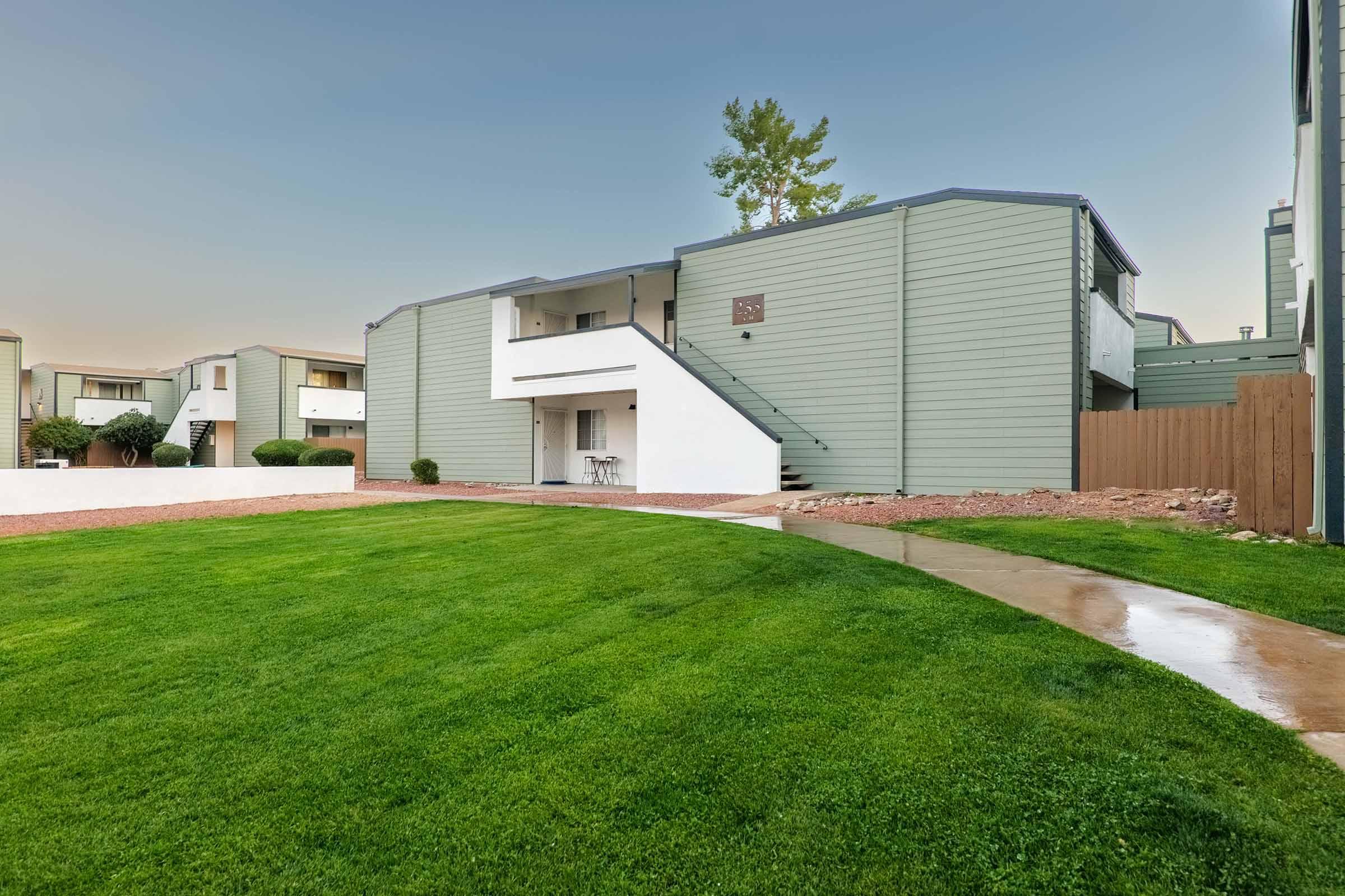A well-maintained multi-unit residential building with light green siding, featuring a set of stairs leading to the second floor. The surrounding area has neatly trimmed grass and a pathway. Nearby, there are small trees and shrubs, adding to the inviting atmosphere of the property.