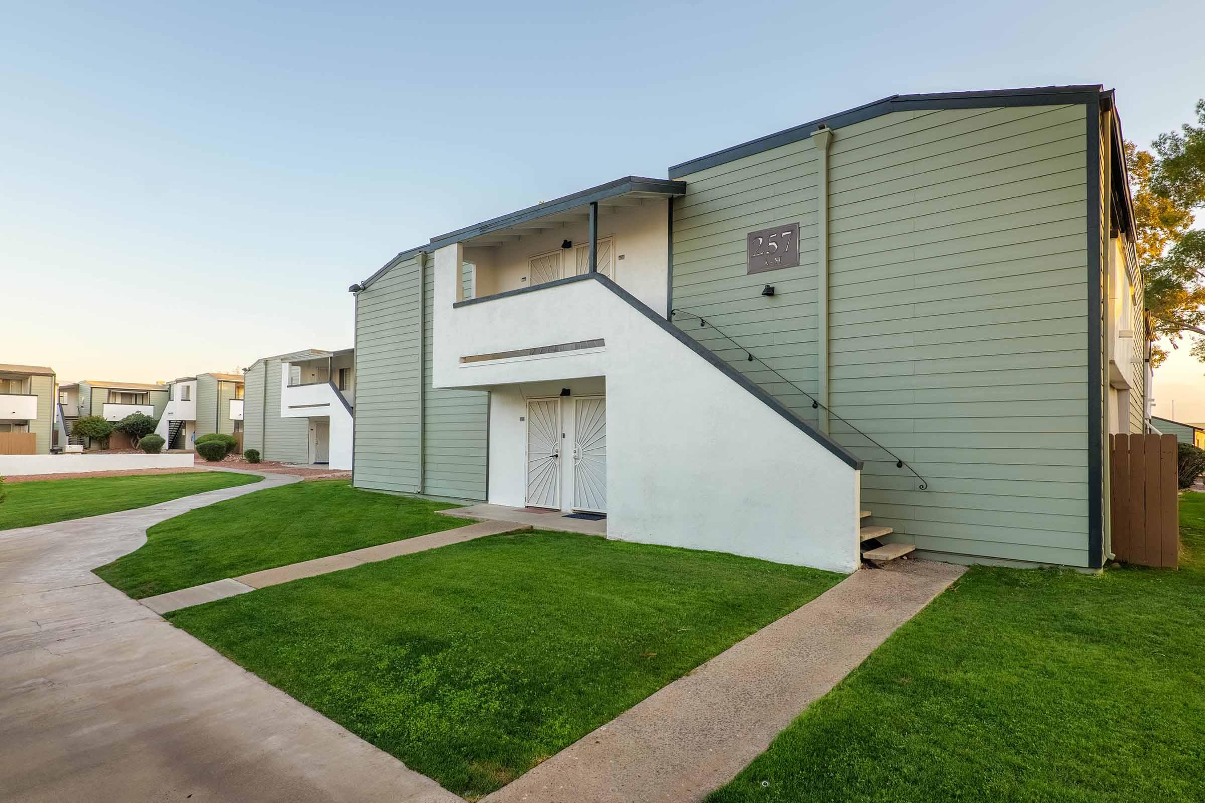 Two-story residential building featuring a light green exterior with white accents. Stairs lead to a second-floor entrance, and a small lawn with neatly kept grass is visible in the foreground. Pathways connect to other nearby buildings in a peaceful setting.