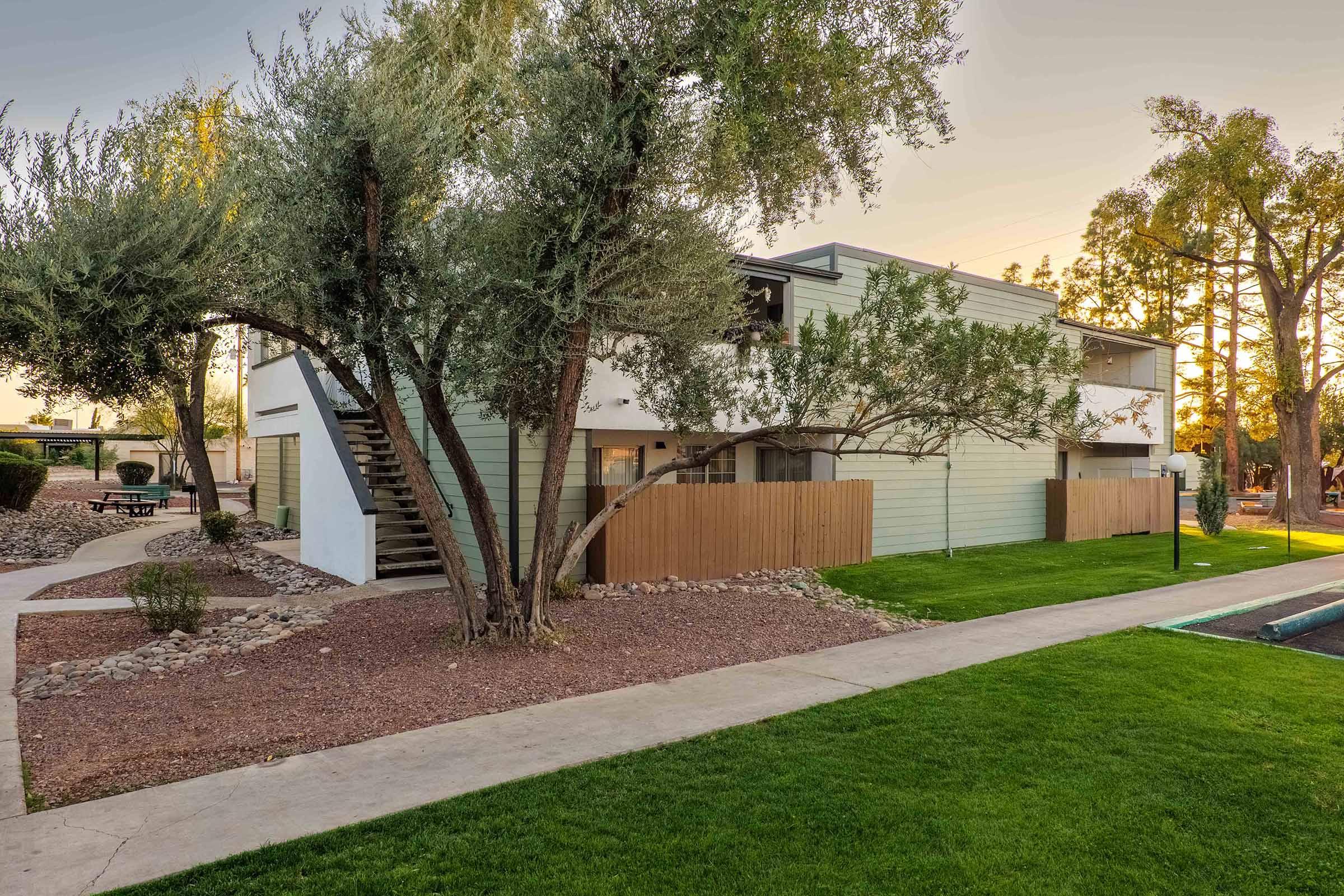 A modern building surrounded by green grass and trees. The structure has a light-colored exterior with a staircase on one side. There is a gravel pathway leading to the entrance and a grassy area nearby, creating a serene outdoor space. The scene is illuminated by warm, evening light.