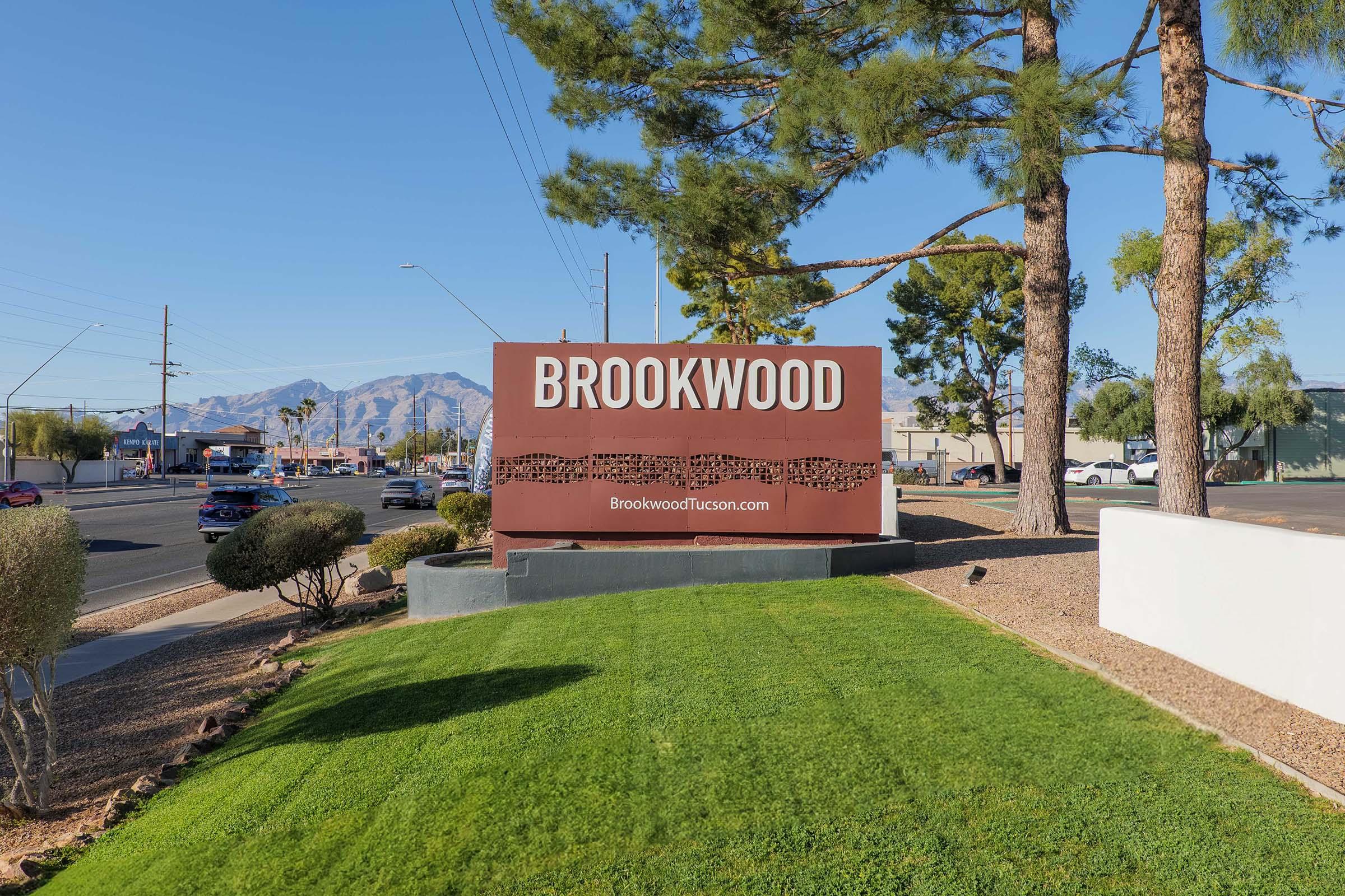 A large sign reading "BROOKWOOD" is visible near a road, surrounded by green grass and desert landscaping. There are a few tall trees in the background, and mountains can be seen in the distance under a clear blue sky. The sign is well-lit and prominent, indicating a location in Tucson.