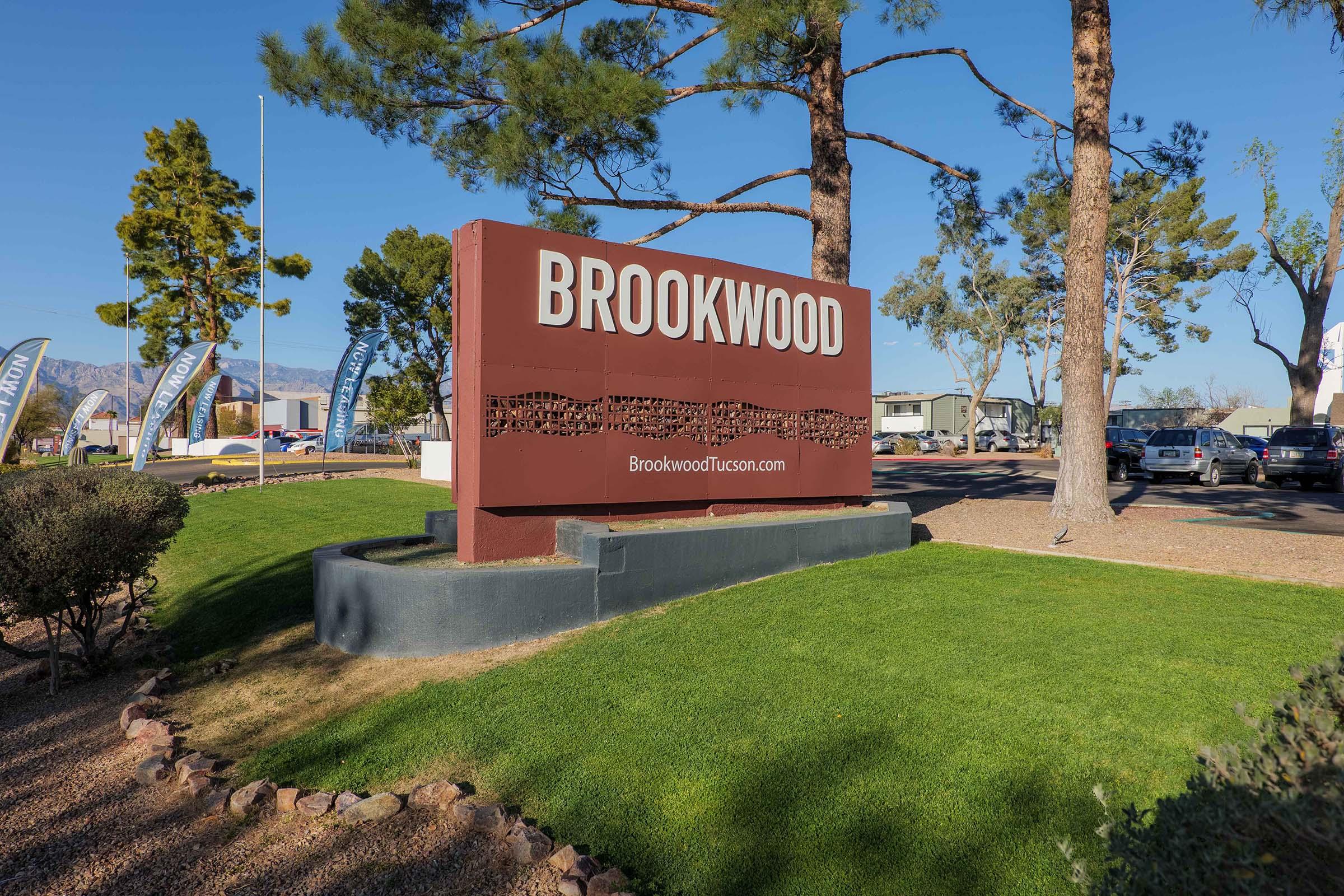 Sign for Brookwood, featuring the name "BROOKWOOD" prominently displayed. Below the name, the website "BrookwoodTucson.com" is visible. The sign is surrounded by well-maintained grass and trees, with a clear blue sky in the background.