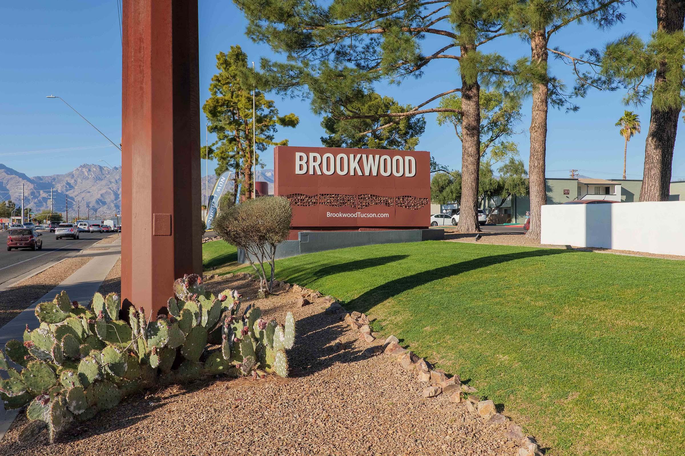 A large sign reading "BROOKWOOD" is prominently displayed at the entrance of a property. Surrounding the sign are green lawns and a landscape featuring palm trees and cacti. The background shows a clear blue sky and distant mountains, with a road visible in front.
