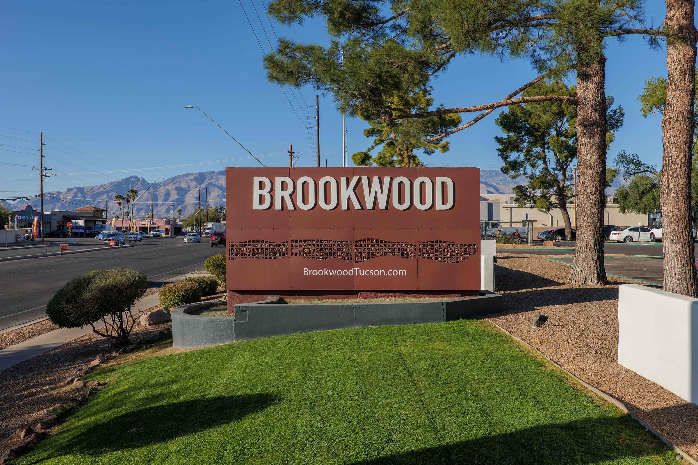 Sign for Brookwood in Tucson, featuring the name "BROOKWOOD" prominently displayed in large letters against a backdrop of mountains. The sign is landscaped with green grass and surrounded by trees, under a clear blue sky. The website "BrookwoodTucson.com" is also visible at the base of the sign.