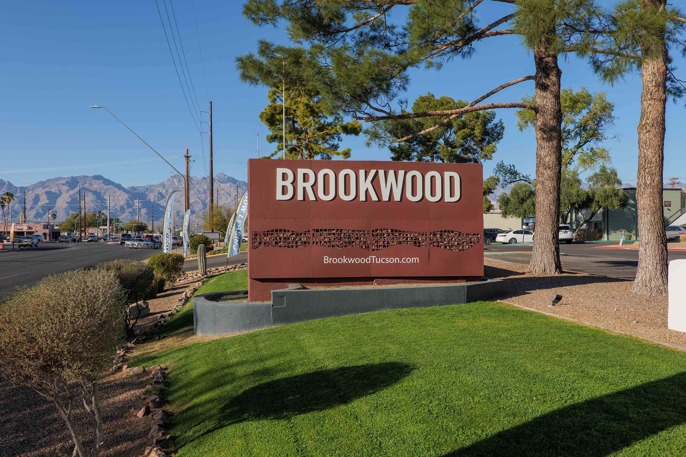 A large outdoor sign featuring the word "BROOKWOOD" prominently displayed. Below the name is the website address "BrookwoodTucson.com." The sign is surrounded by grass and small shrubs, with mountains and a clear blue sky in the background. Power lines are visible along the road leading up to the sign.