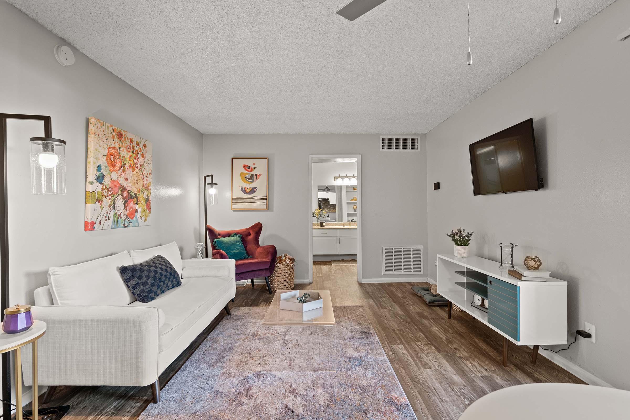 A cozy living room featuring a white sofa with decorative cushions, a colorful floral wall art, and a rustic wooden coffee table. There is a plush armchair in deep red, a flat-screen TV mounted on the wall, and a light wood floor. A glimpse of a modern kitchen is visible in the background.