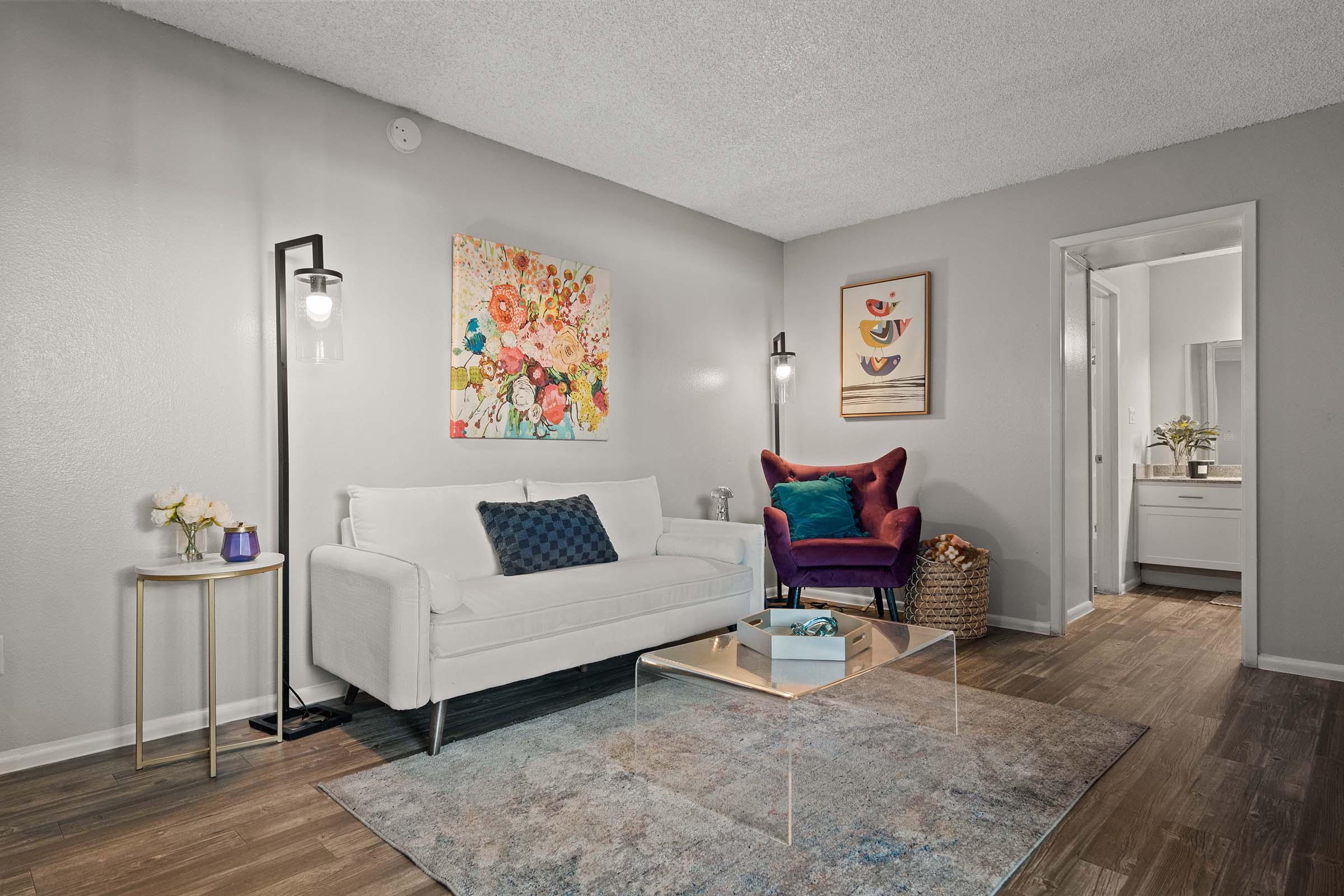 A cozy living room featuring a white sofa with decorative pillows, a purple accent chair, and a small side table with flowers. Colorful floral artwork adorns the wall, and a light wooden floor complements the neutral grey walls. A doorway leads to a bright, modern bathroom. Soft lighting creates a welcoming atmosphere.