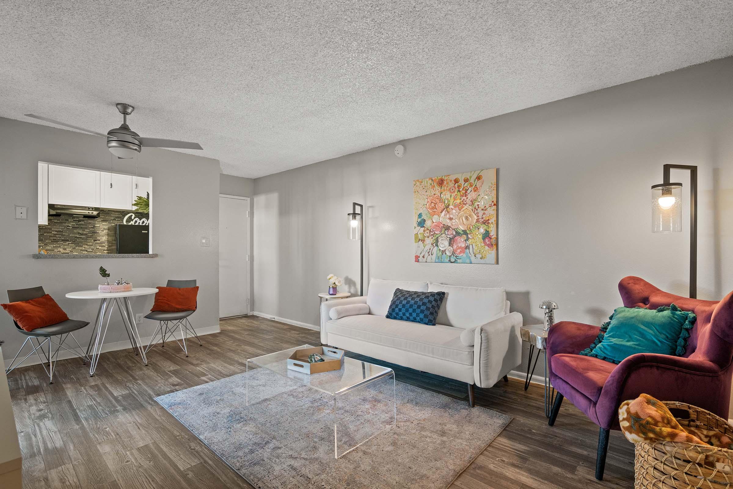 Cozy living room featuring a white sofa, a purple chair, and a glass coffee table. There is a small dining area with a round table and four chairs. The walls are light gray, decorated with a floral painting. A ceiling fan is visible, and the space is bright and inviting.