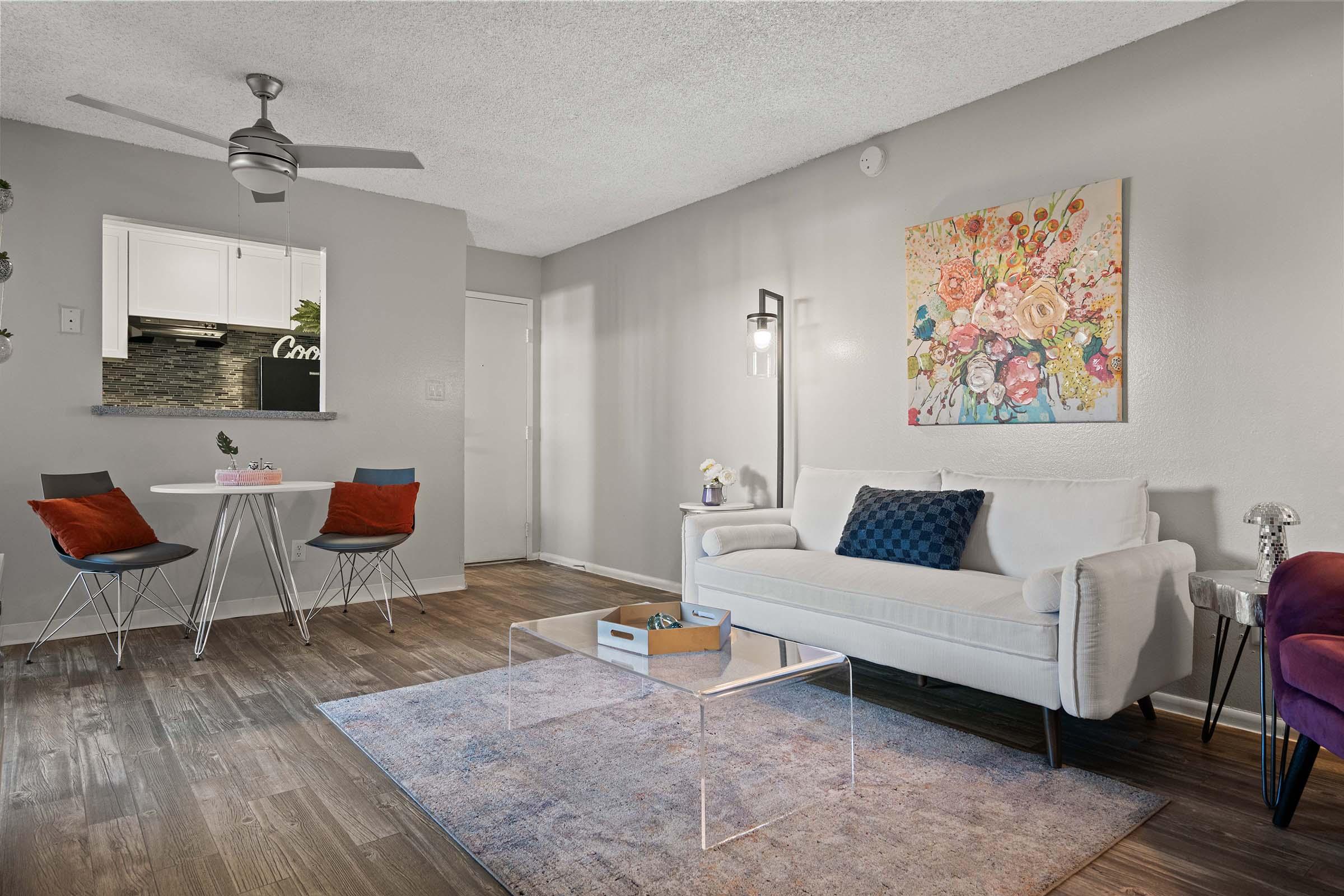 A cozy living room featuring a white sofa with decorative cushions, a glass coffee table on a rug, and a floral wall art piece. A small dining area with a round table and colorful chairs is visible. The space is well-lit with a ceiling fan and modern decor, creating a welcoming atmosphere.