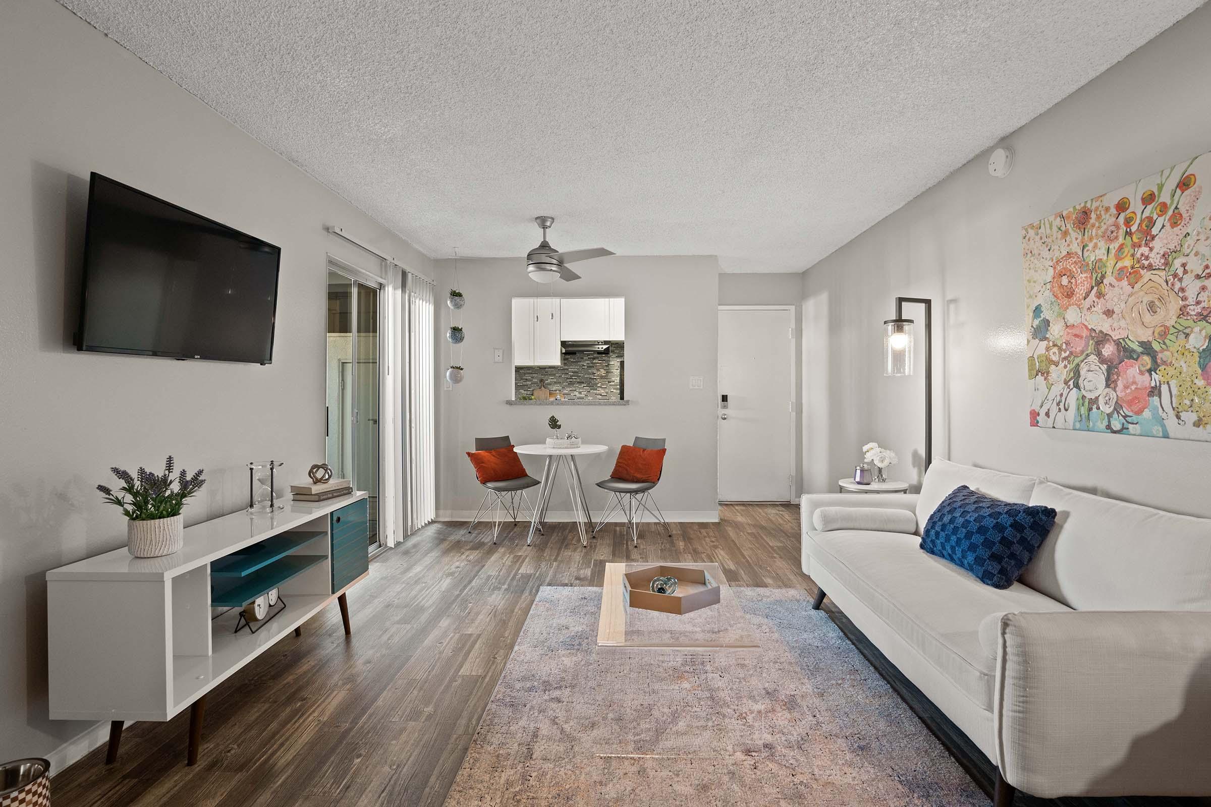 A cozy living room featuring a white sofa with blue cushions, a wooden coffee table, and a wall-mounted TV. There is a small dining area with a round table and two chairs. The room has light gray walls, wooden flooring, and colorful floral artwork, providing a warm and inviting atmosphere. A ceiling fan is also visible.