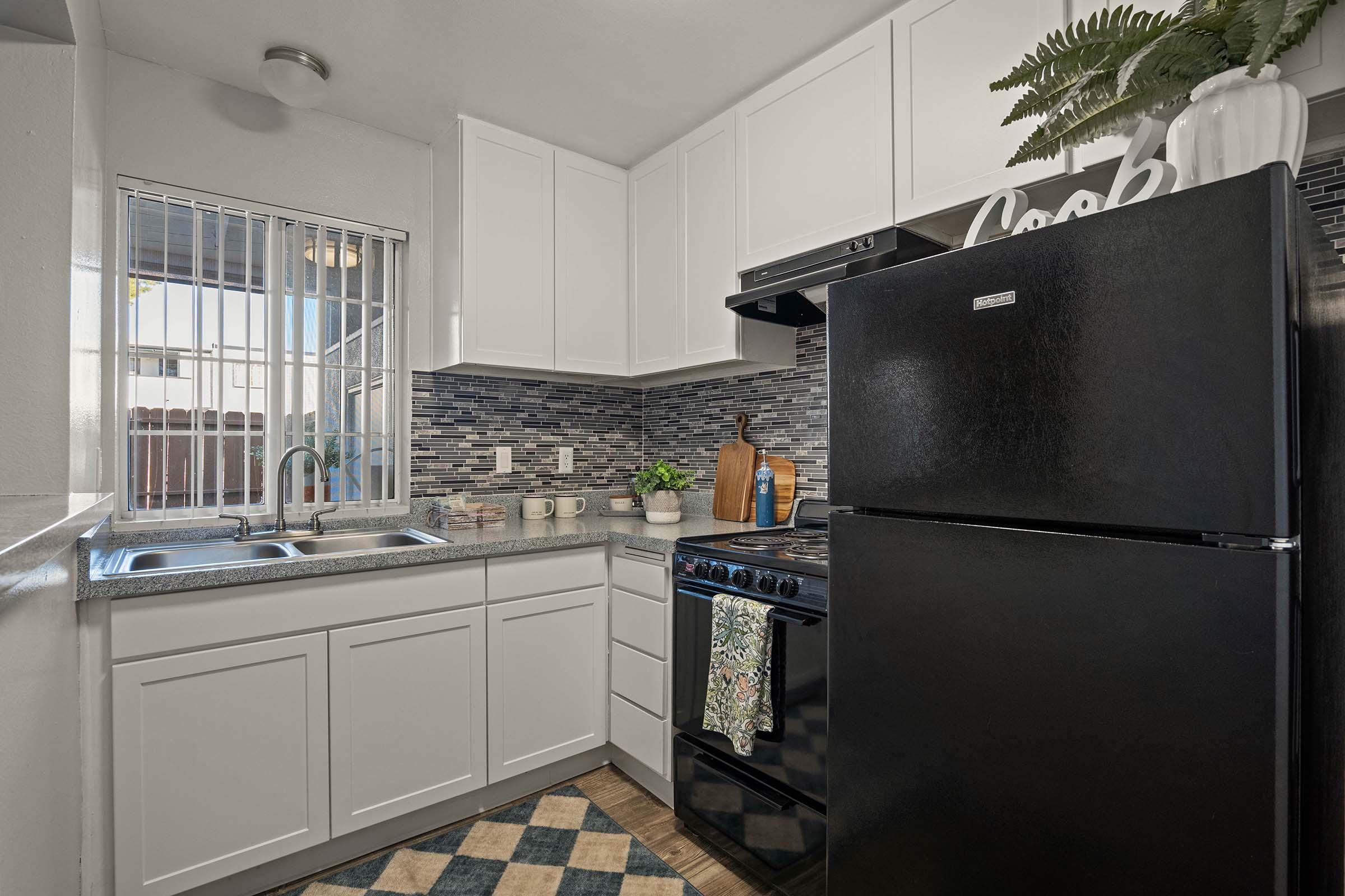 A modern kitchen featuring white cabinetry, a black refrigerator, and a black stove. There is a sink under a window with blinds, and a countertop displaying decorative items like a wooden cutting board and plants. The backsplash has blue and gray tiles, and a patterned rug is on the floor.
