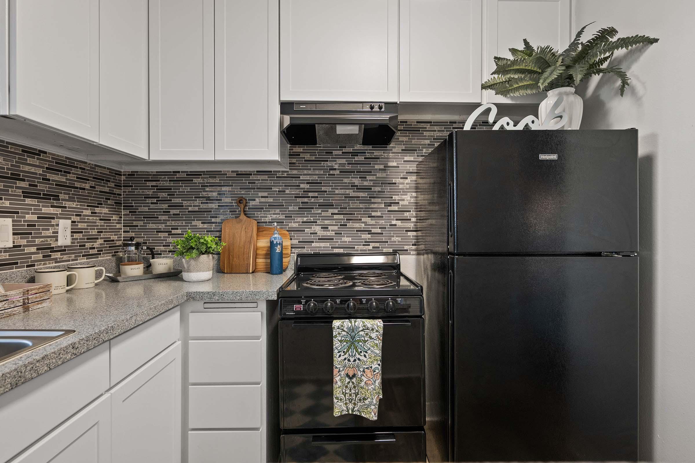 A modern kitchen with white cabinets, a black refrigerator, and a black stove. The countertop features a cutting board, small potted plants, and utensils. A decorative towel hangs on the stove, and a stylish plant sits on the shelf above. The backsplash is made of patterned tiles in shades of gray and black.