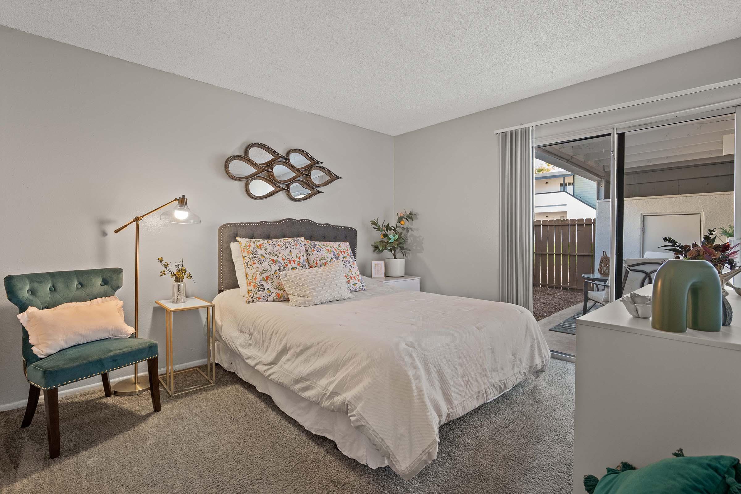 A cozy bedroom featuring a queen-sized bed with decorative pillows, a stylish wall mirror, and a bedside lamp. A green accent chair sits next to the bed, and large sliding glass doors open to a small outdoor space with patio seating. The walls are painted in soft neutral tones.